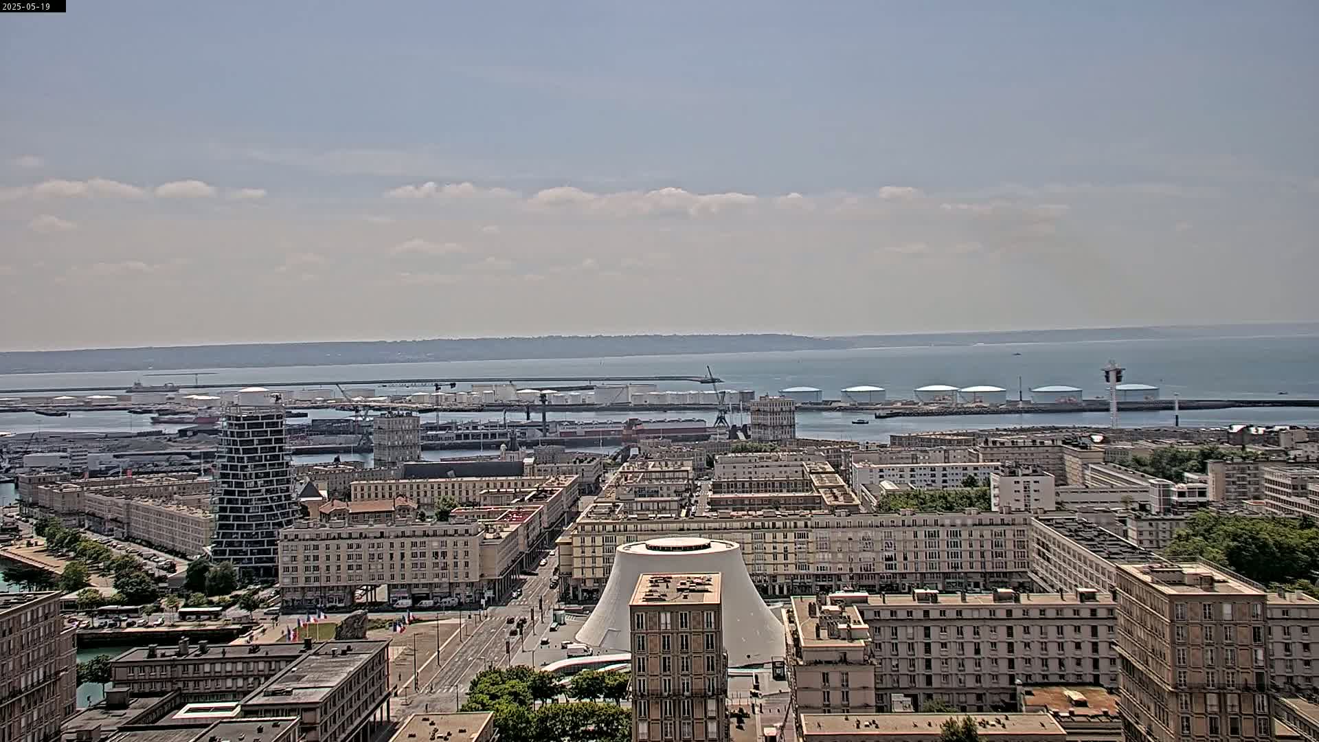 A partly cloudy day overlooks a city harbor with numerous buildings, a large body of water, and industrial storage tanks.