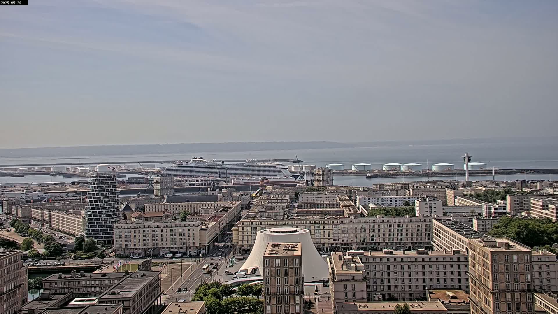 A hazy day reveals a cityscape with a large cruise ship docked near an industrial area, all overlooking a calm body of water.