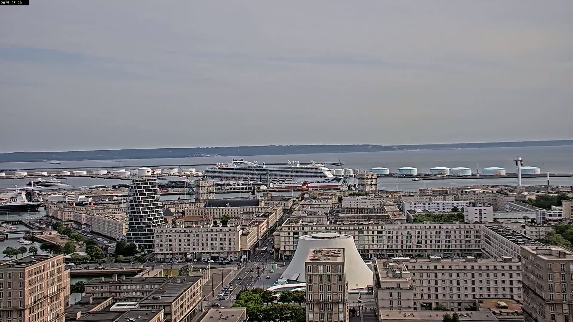 An aerial view of a city harbor on a mostly cloudy day shows a large cruise ship docked near several industrial buildings and numerous apartment buildings.