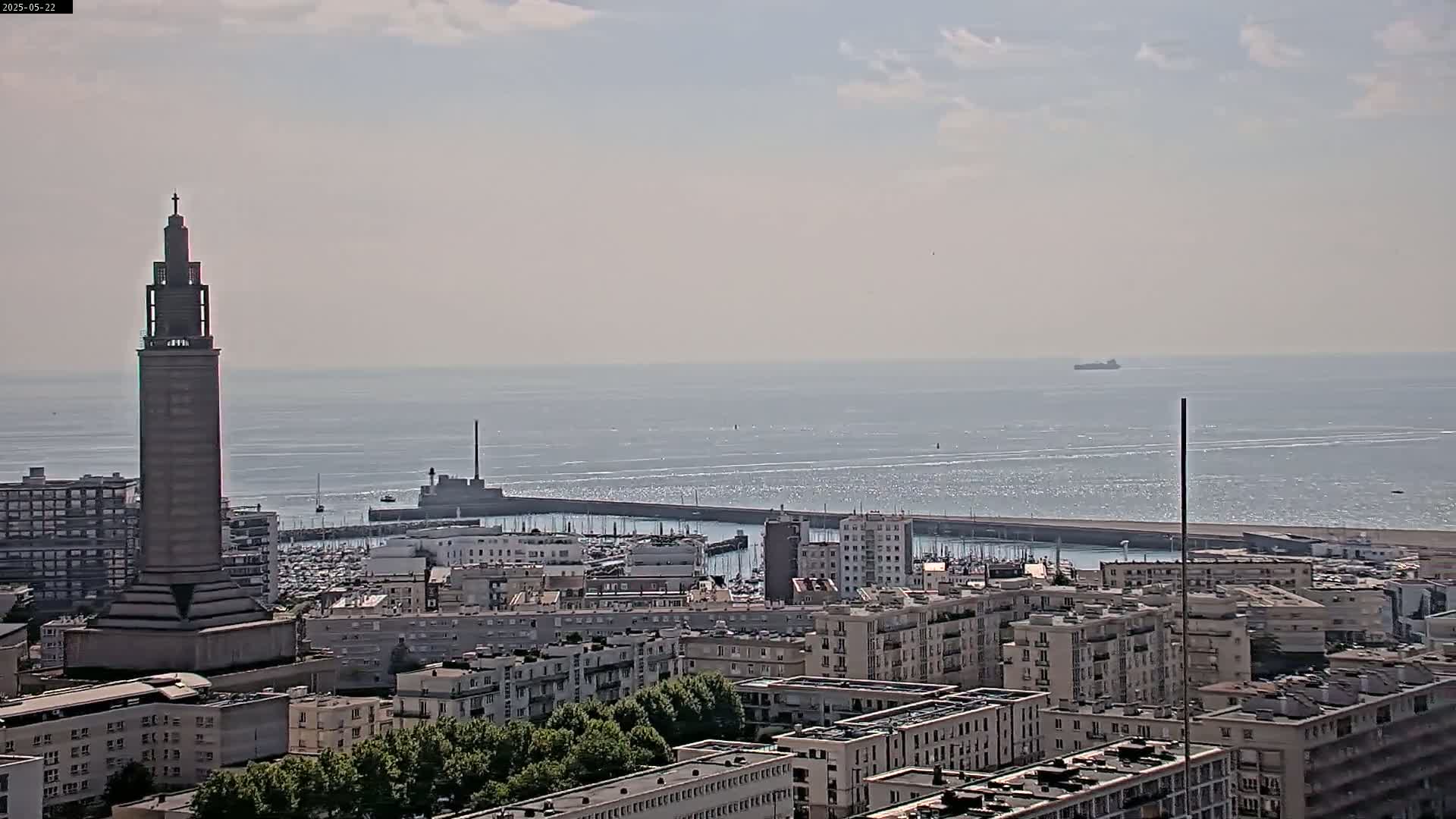A tall tower stands prominently over a coastal city with a harbor filled with boats under a hazy, sunny sky, and a large body of water extends to the horizon.