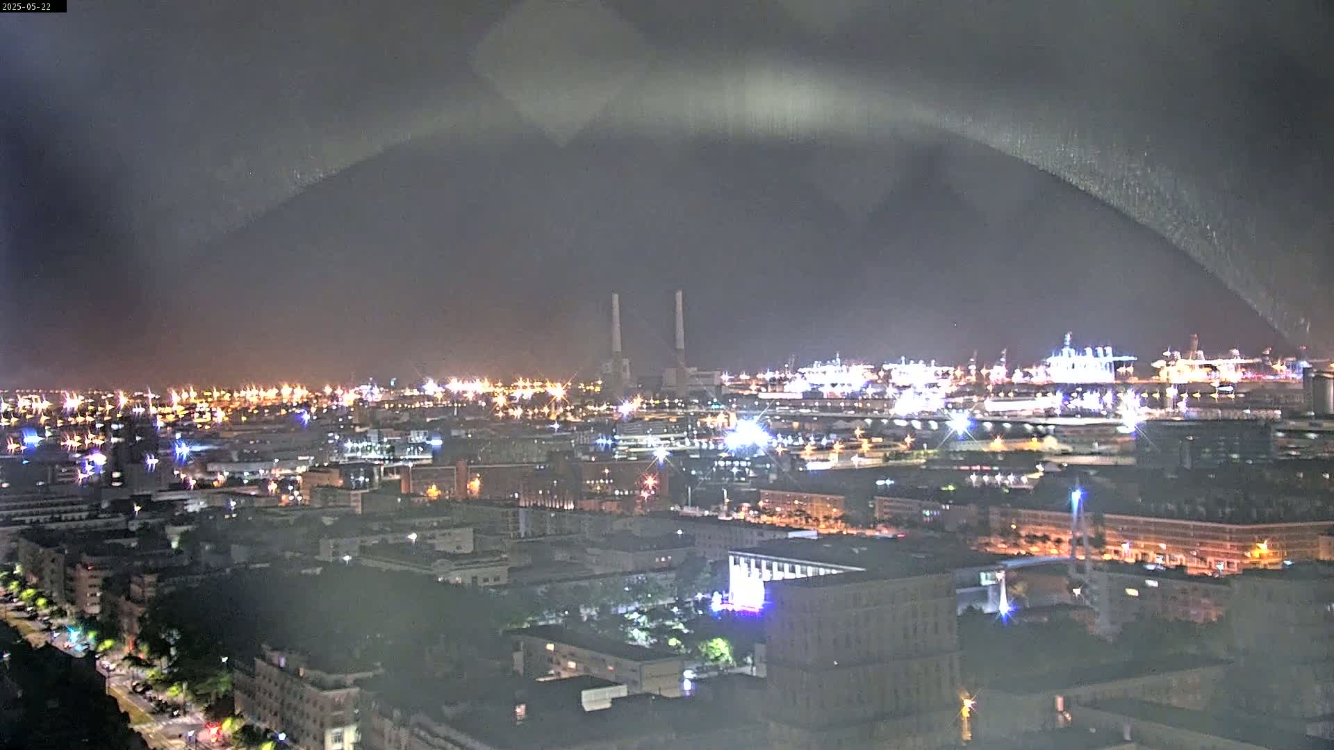 A nighttime view of a city harbor with illuminated buildings and industrial areas under a dark, cloudy sky.