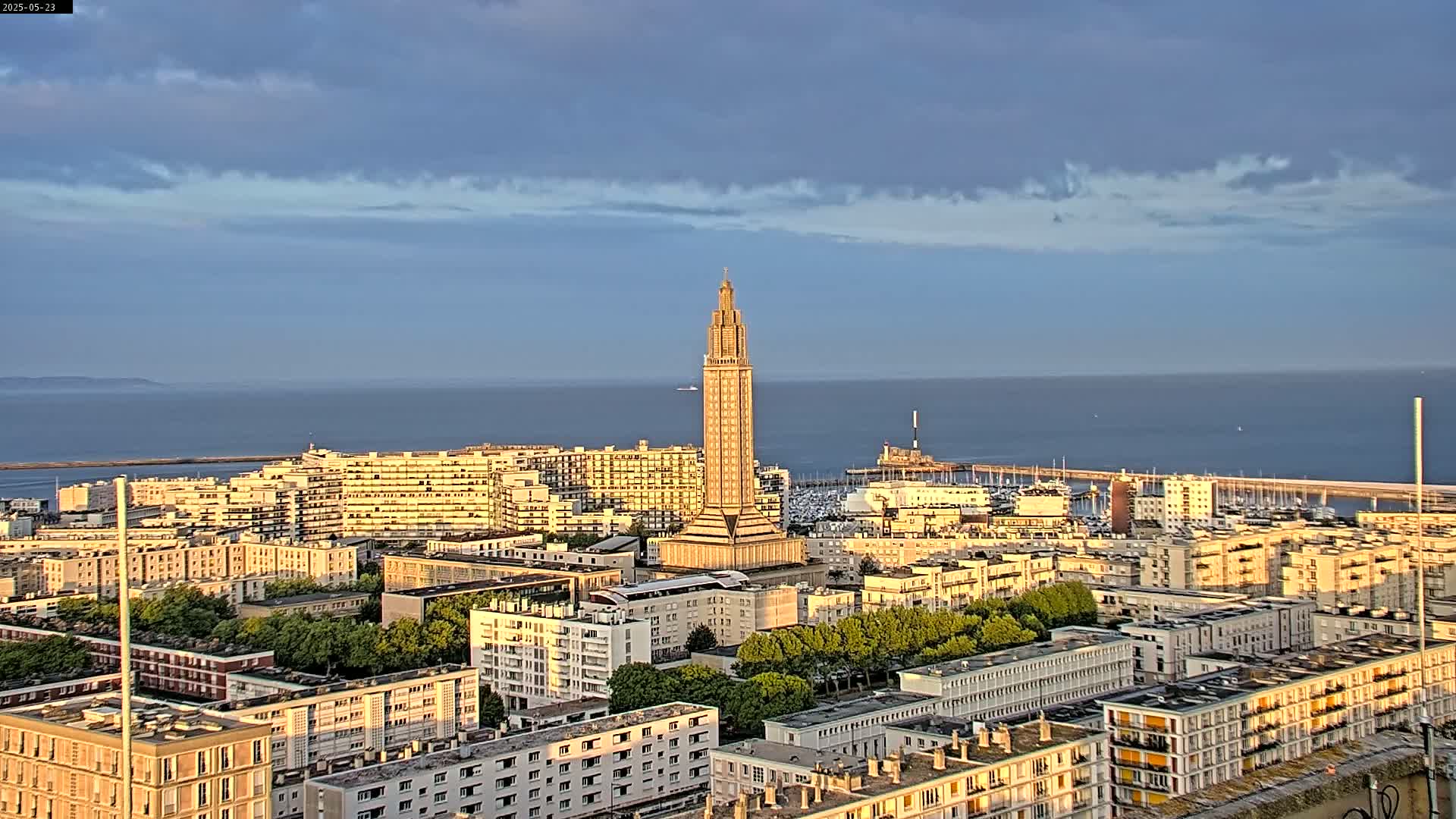 A tall, slender tower stands in the center of a coastal city under a partly cloudy sky, overlooking a calm body of water with a harbor filled with boats.