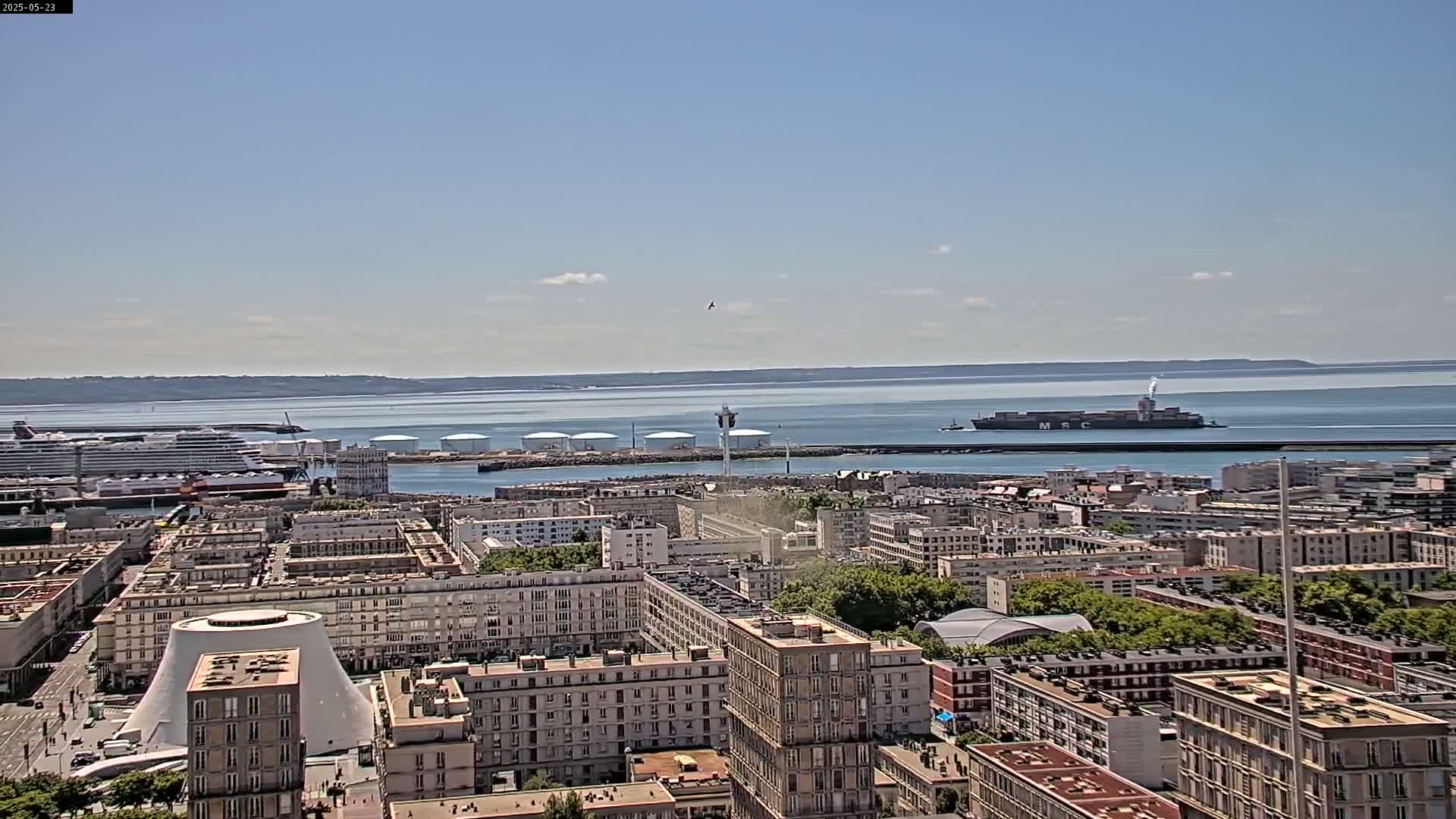 A high-angle view shows a city harbor on a sunny day, with several ships including a large cruise ship and a container ship visible in the water, and numerous buildings extending into the background.