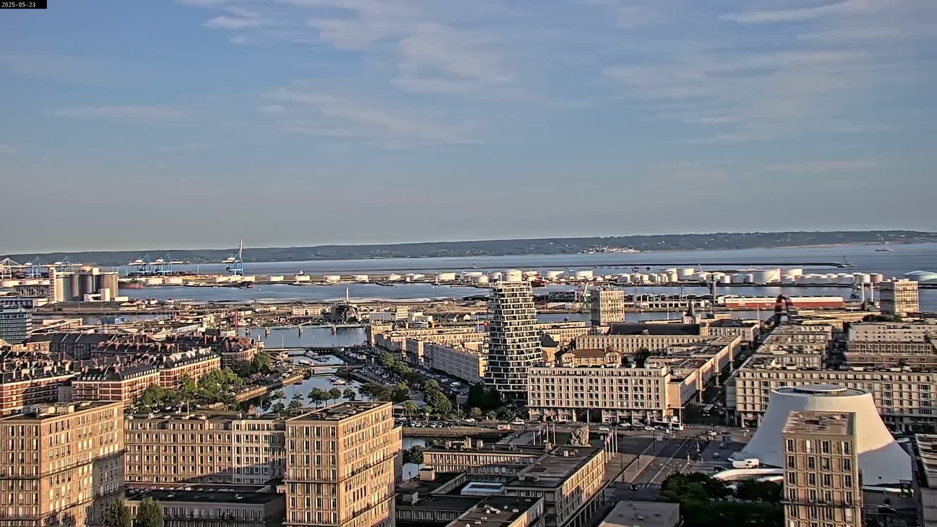 A sprawling cityscape with numerous buildings and a large body of water in the background under a mostly clear sky.