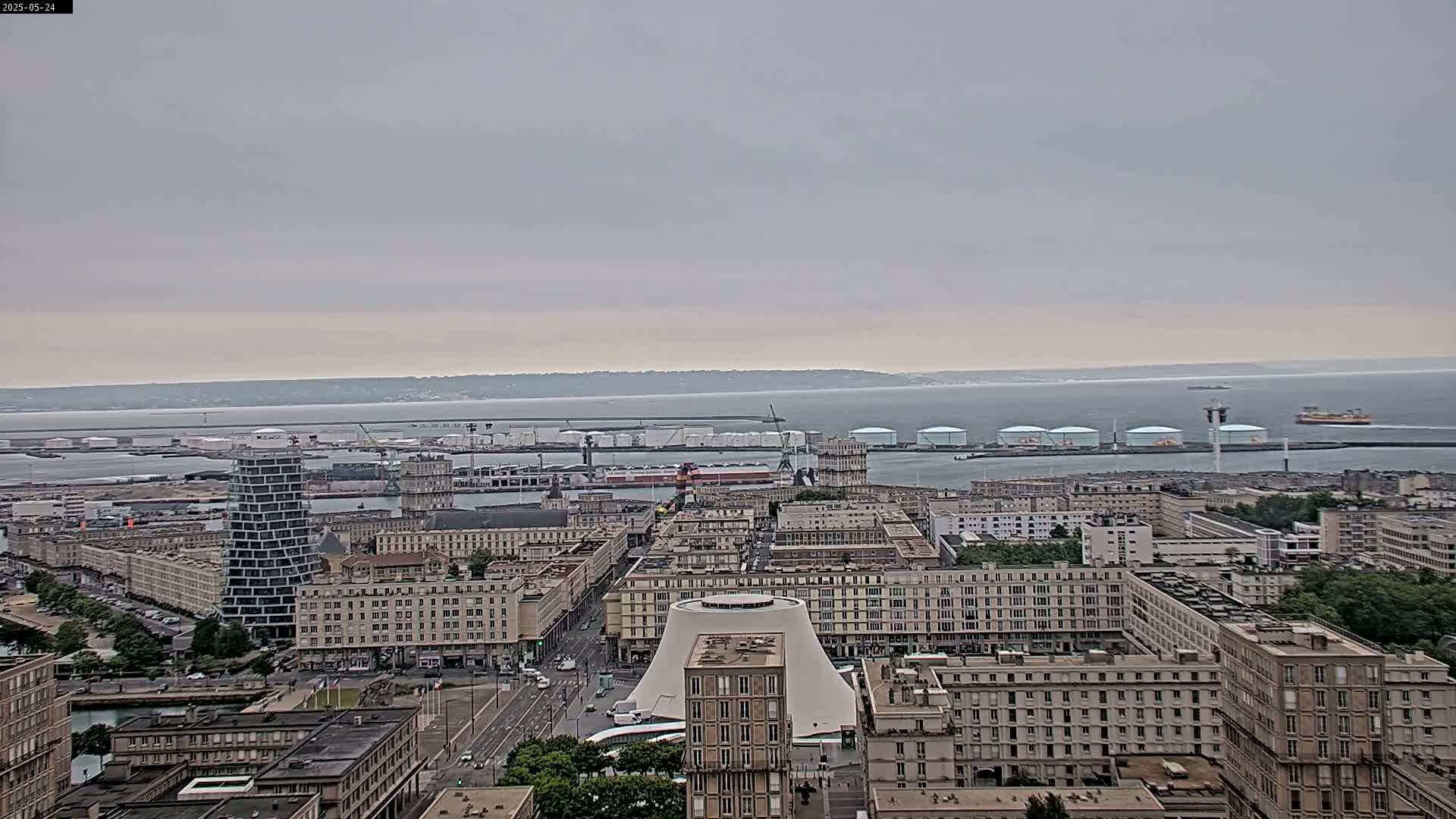 A high-angle, long shot shows a cityscape on an overcast day, with a body of water and a distant horizon in the background.