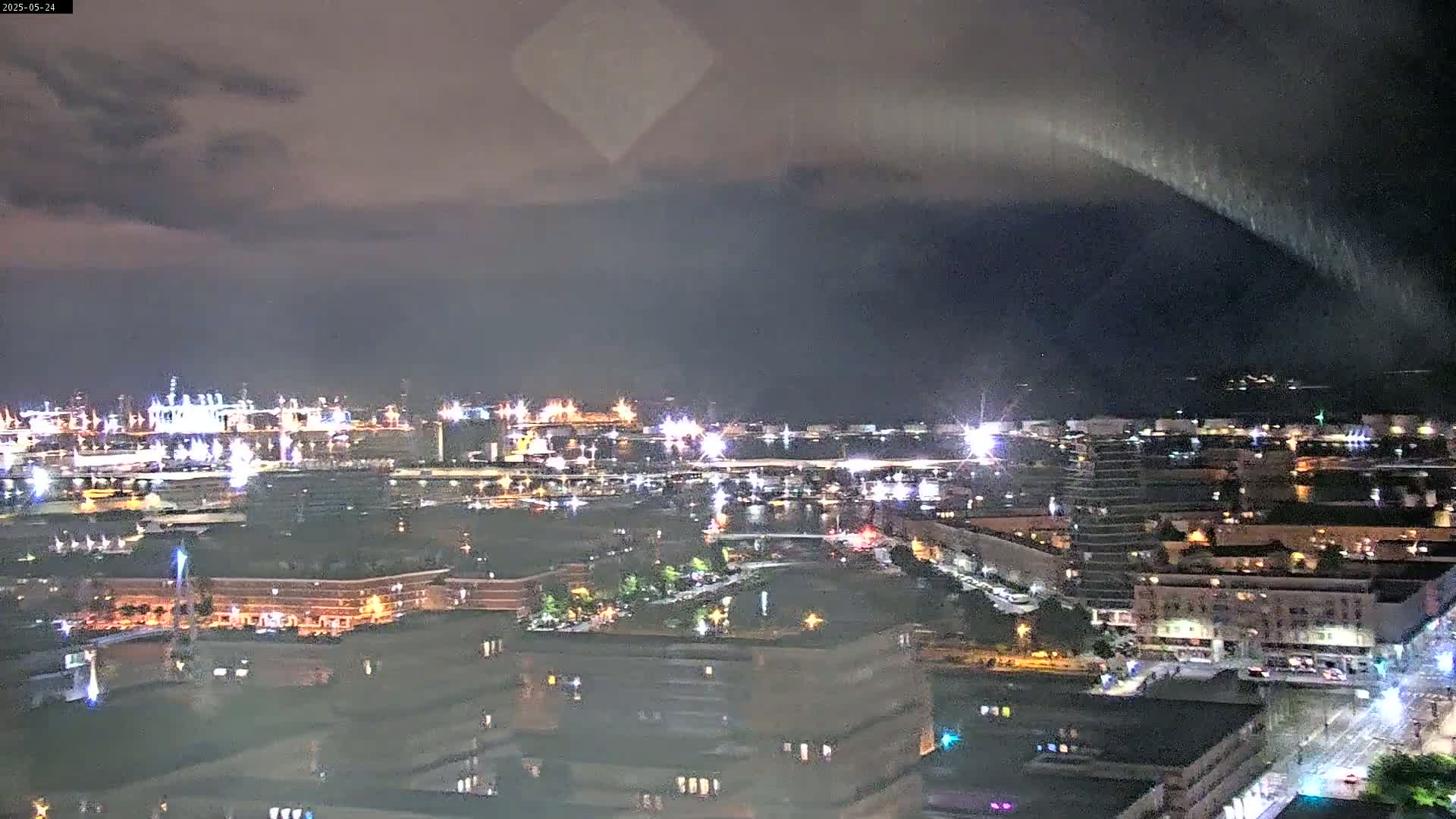A nighttime view of a harbor city under a cloudy sky shows a vast industrial area with many lights, along with buildings and streets.