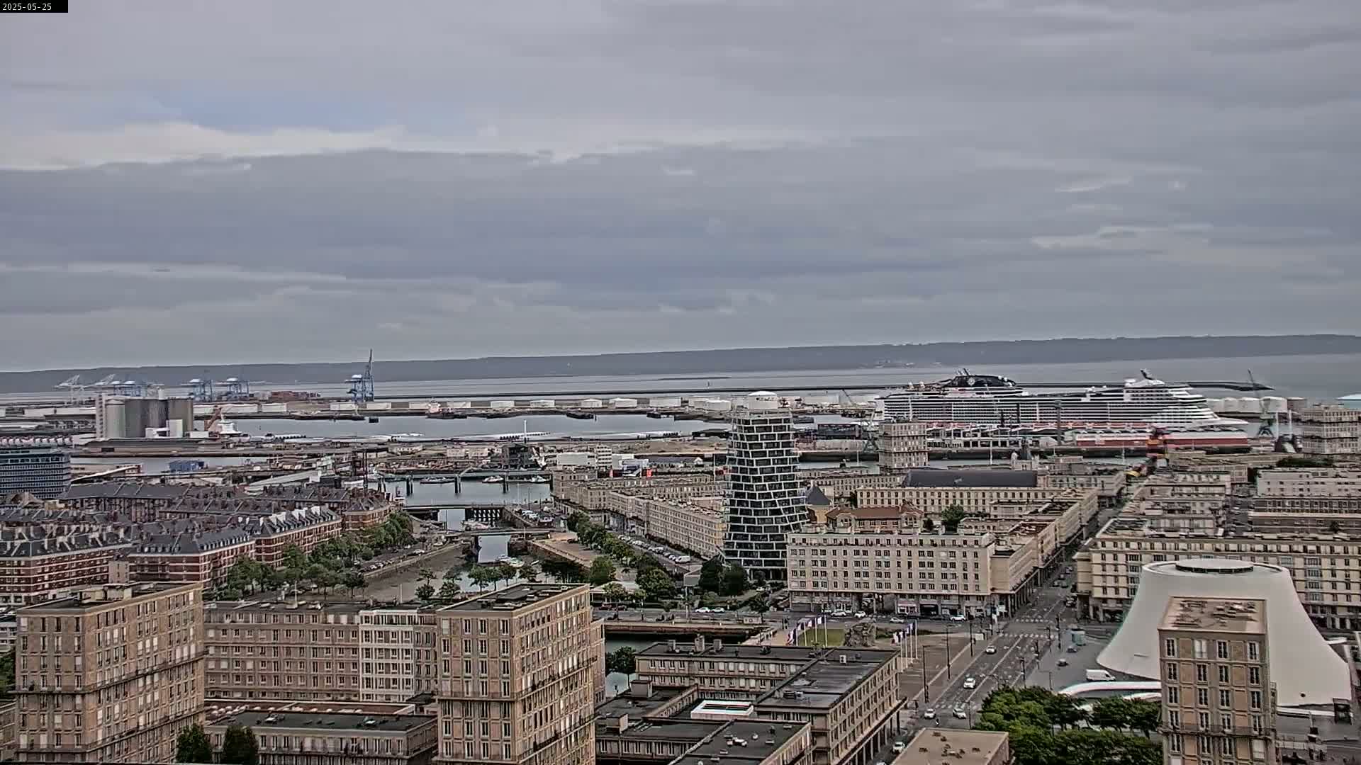 A high-angle view of a city harbor on an overcast day shows a large cruise ship docked near a busy port with numerous buildings and a body of water extending to the horizon.