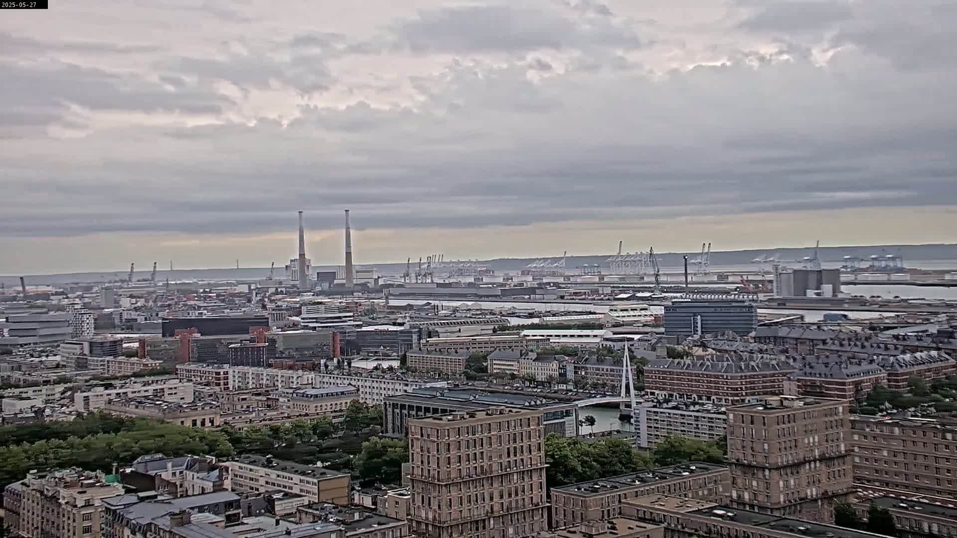 A high-angle view of a city under a cloudy sky shows a large industrial port area in the distance with numerous cranes, and a closer view of several buildings with green trees and waterways.