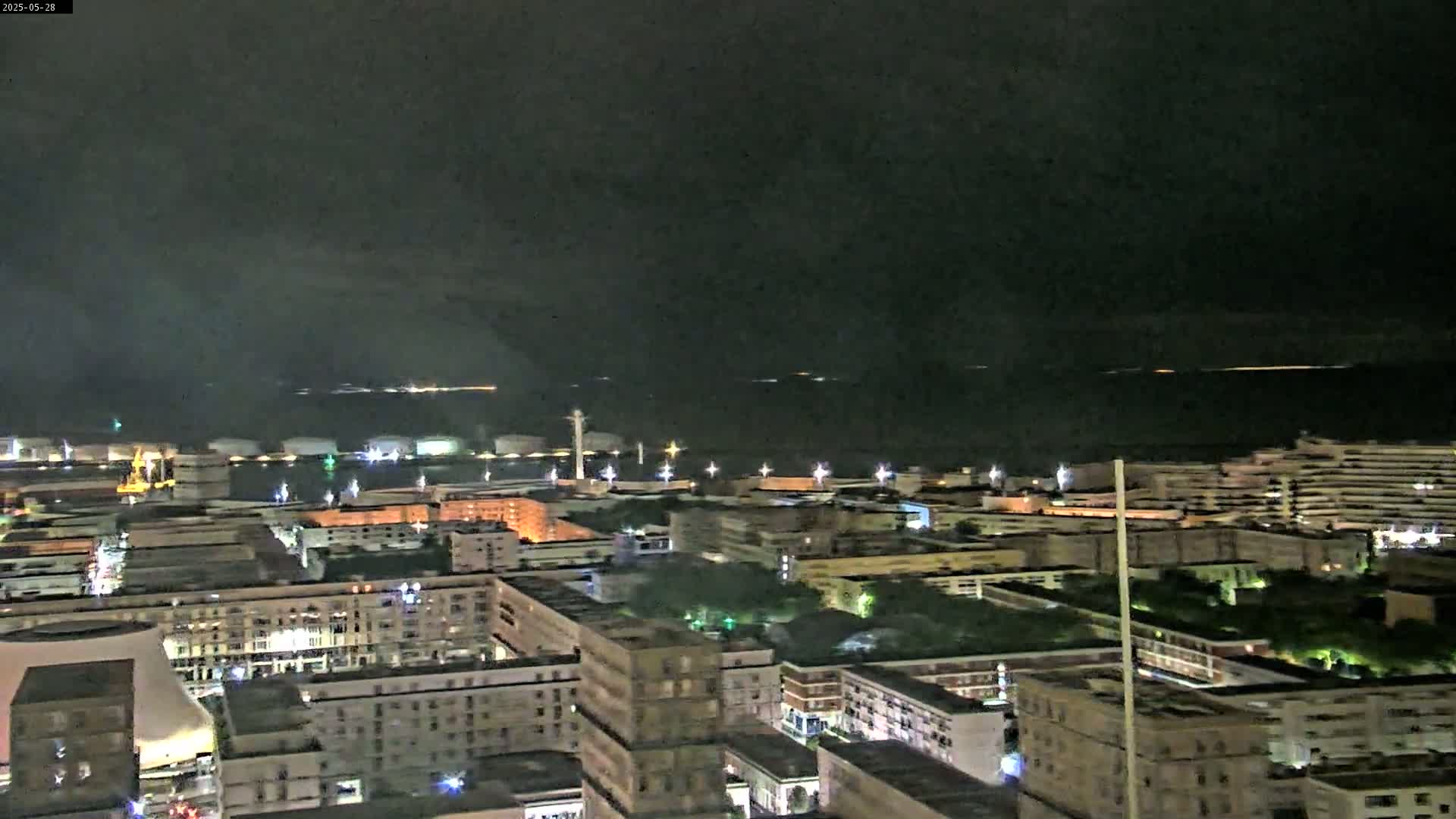 A nighttime, high-angle view of a city's harbor and residential areas, with a calm body of water in the background under a dark sky.