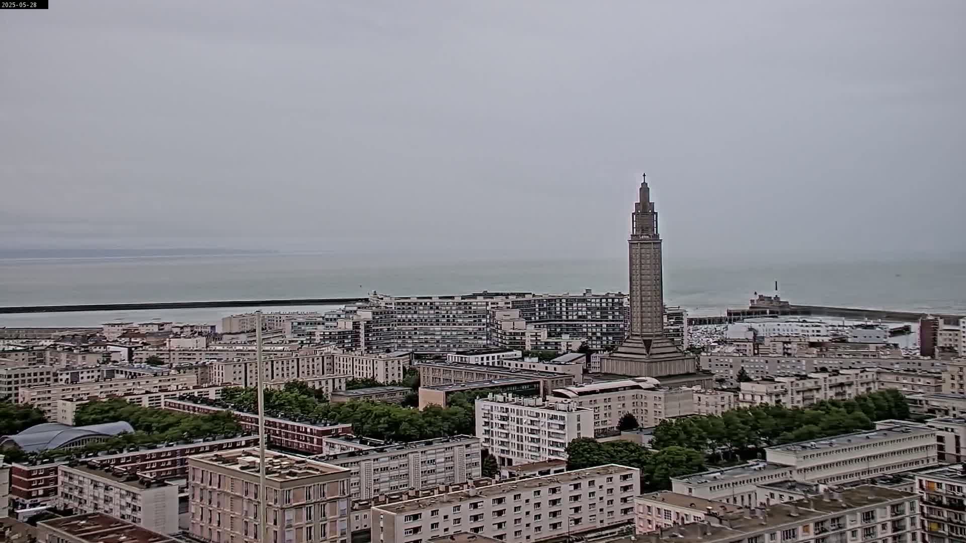 A coastal city with a tall, slender tower is shown on an overcast day, with a calm sea visible in the distance.
