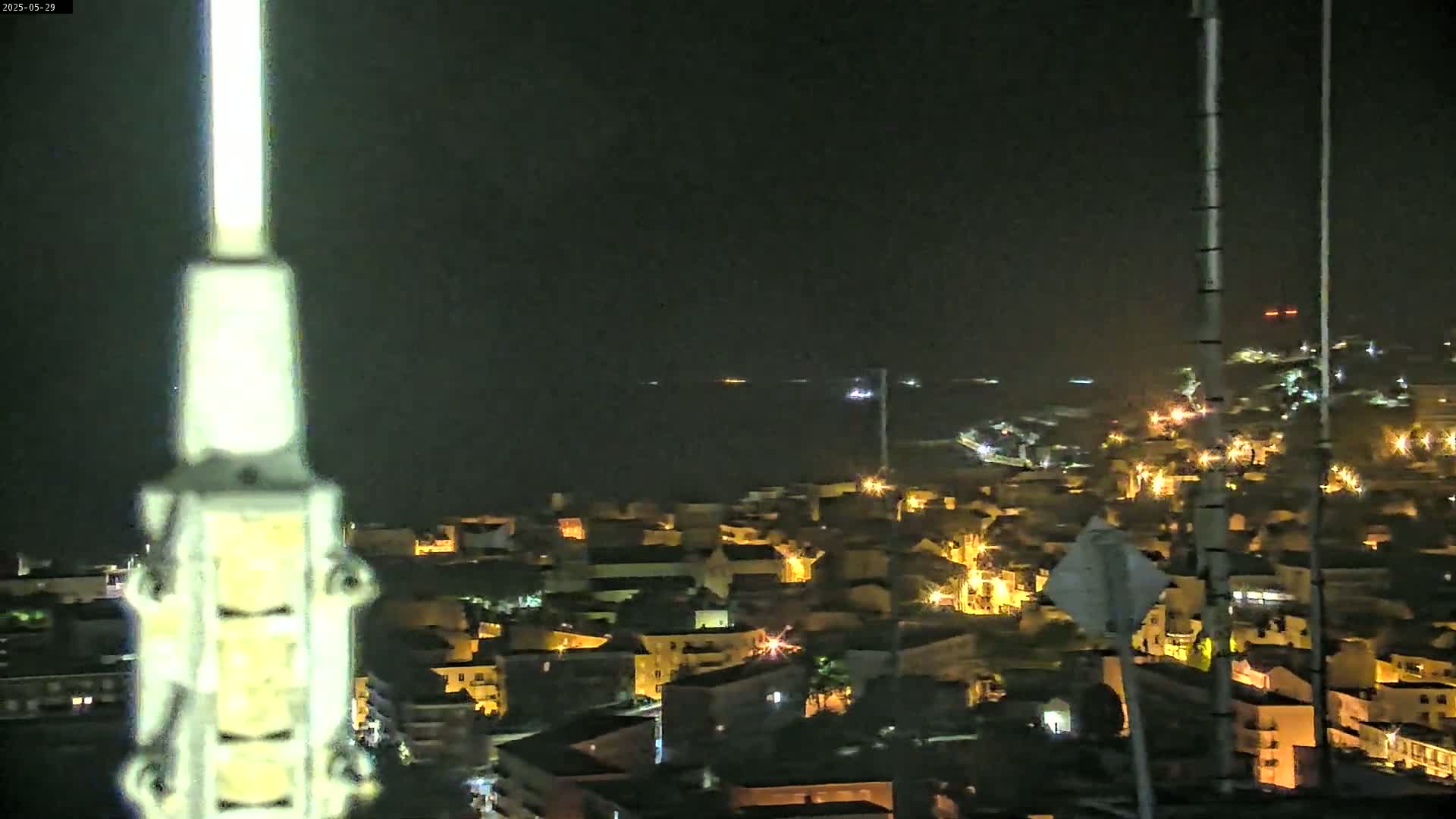 A nighttime, high-angle view shows a coastal town illuminated by streetlights, with a dark body of water visible in the distance under a clear, dark sky.