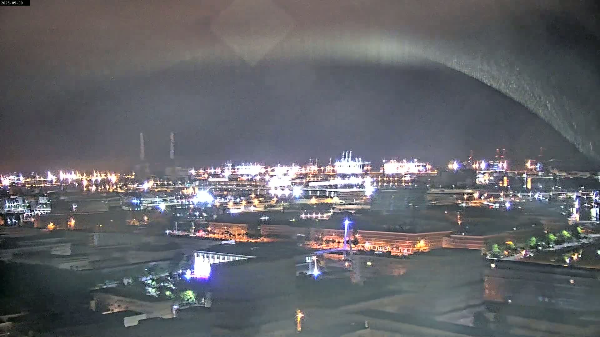A nighttime, possibly rainy, view of an industrial waterfront area with numerous lights illuminating buildings and structures, under a dark, cloudy sky.