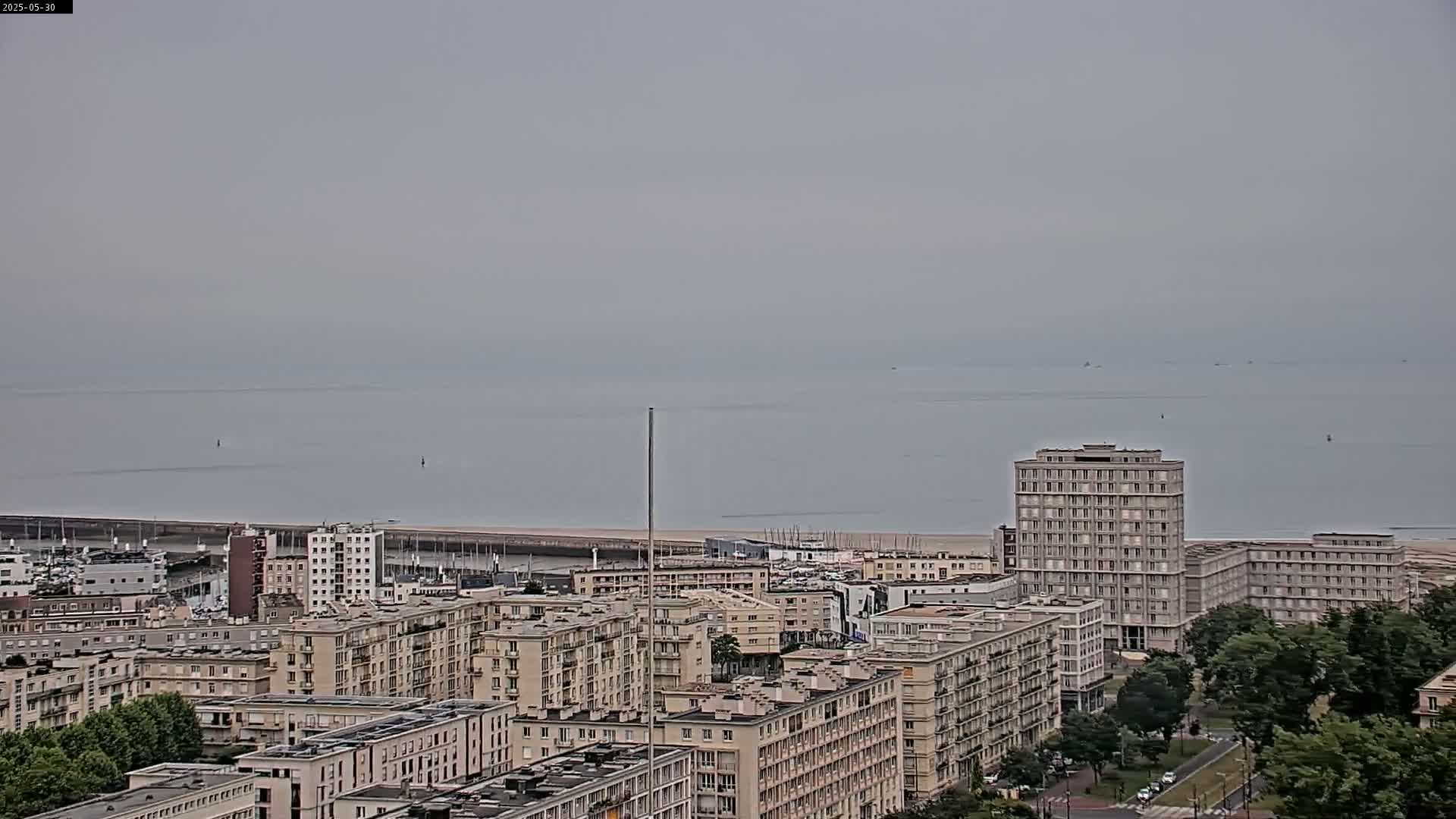 A calm, overcast day reveals a coastal city with numerous apartment buildings, a marina, and a body of water extending to the horizon.