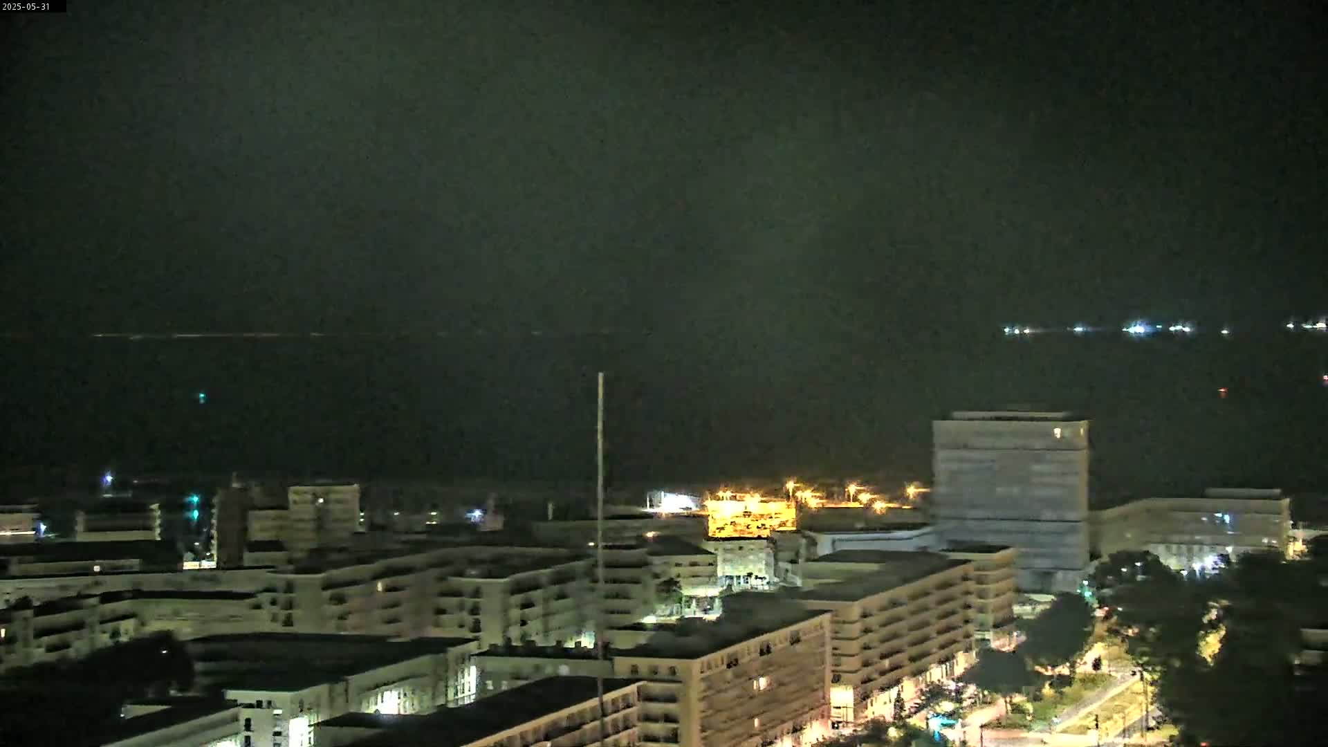 A nighttime, high-angle view shows a city skyline with multi-story buildings illuminated against a dark, overcast sky, overlooking a body of water with faint lights visible in the distance.