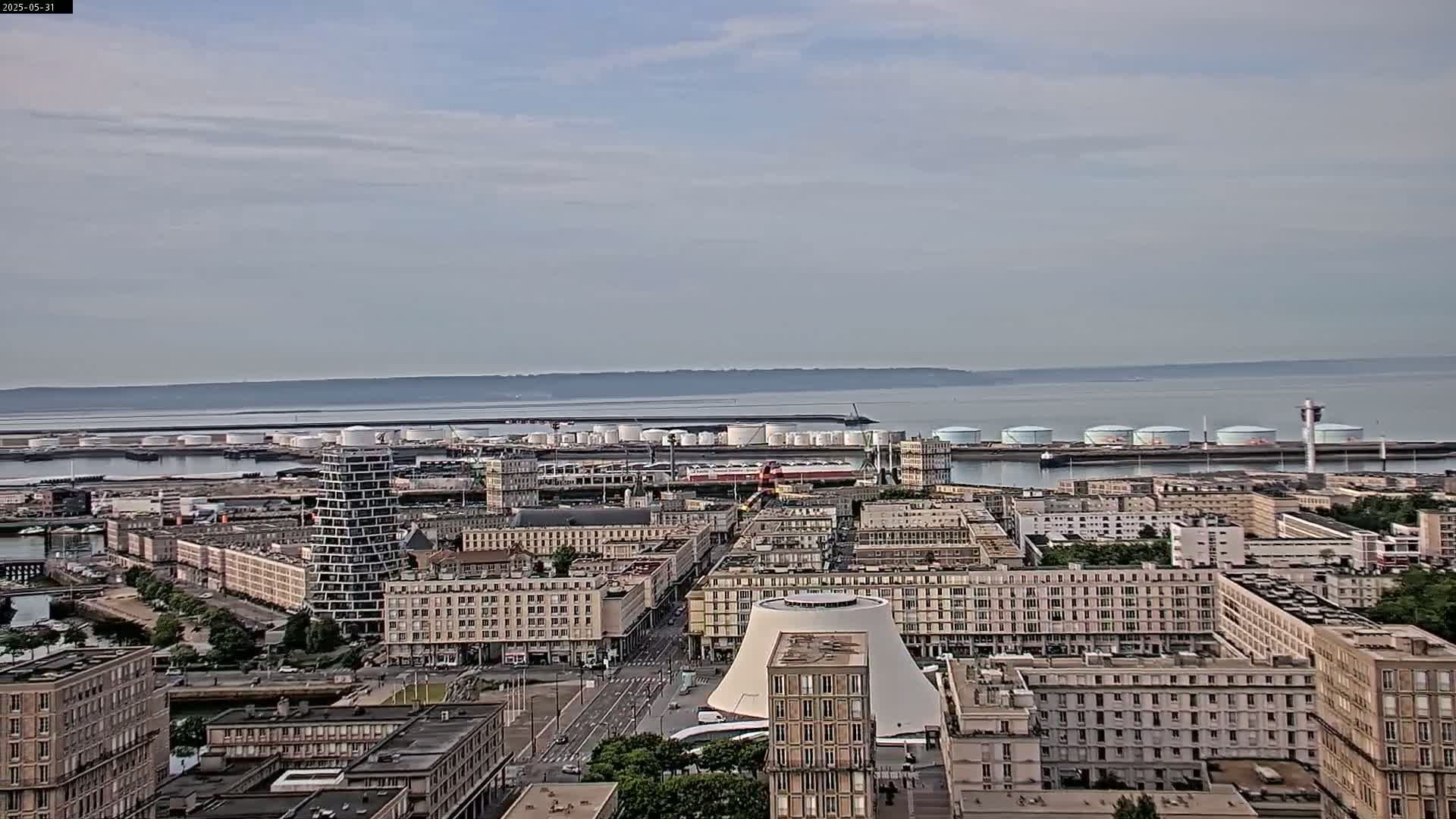 An aerial view shows a city with numerous buildings under a mostly cloudy sky, overlooking a harbor with large storage tanks and a calm body of water beyond.