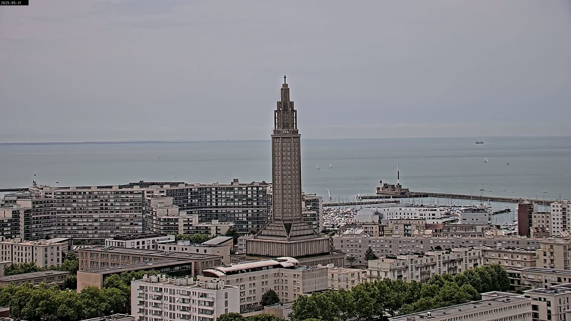 A tall, slender tower stands prominently in a cityscape overlooking a calm sea on an overcast day.