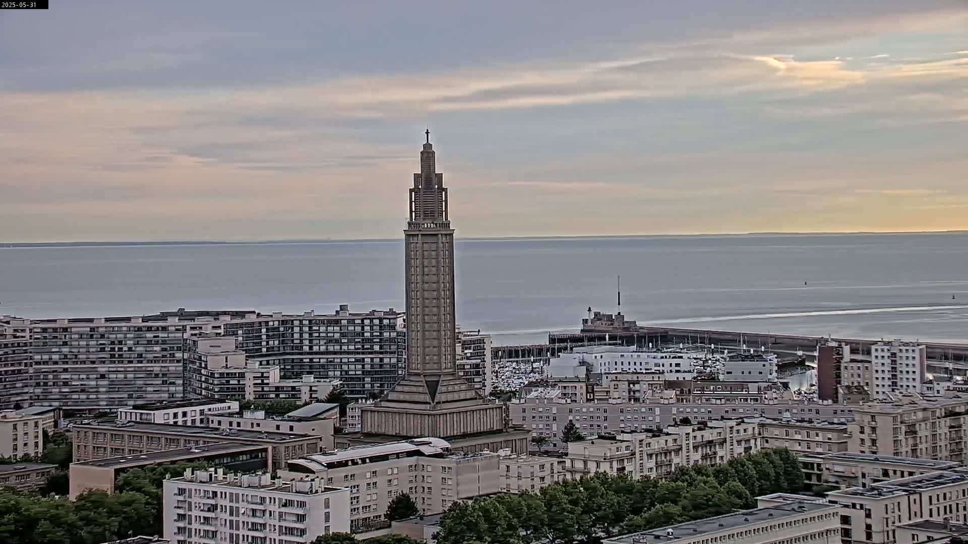 A tall, slender tower stands in the center of a cityscape overlooking a calm sea under a partly cloudy sky.