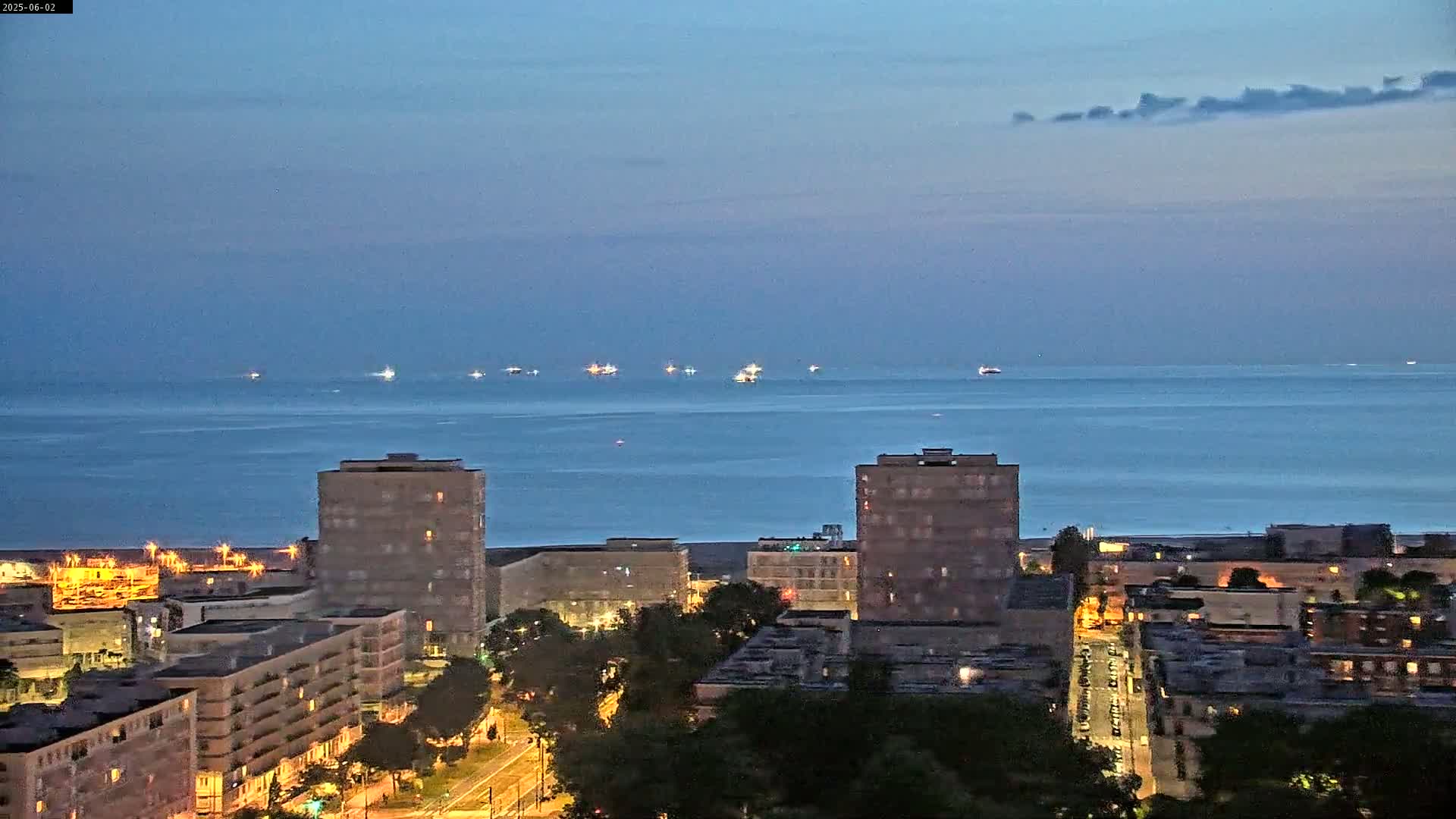 A twilight view shows a calm body of water with several ships in the distance, beyond a city of illuminated buildings and trees.