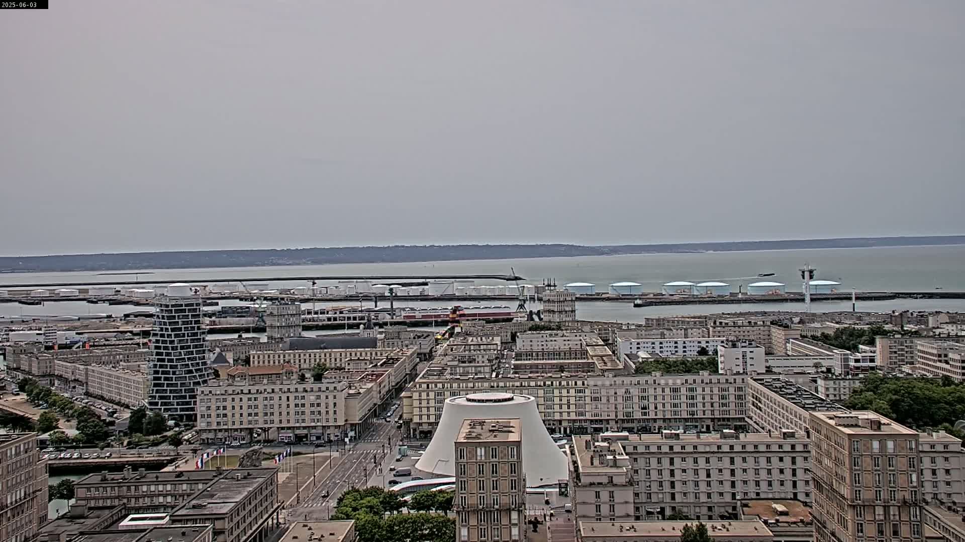 A high-angle, long shot shows a city's harbor with many buildings, including a large, dome-shaped structure, on an overcast day.