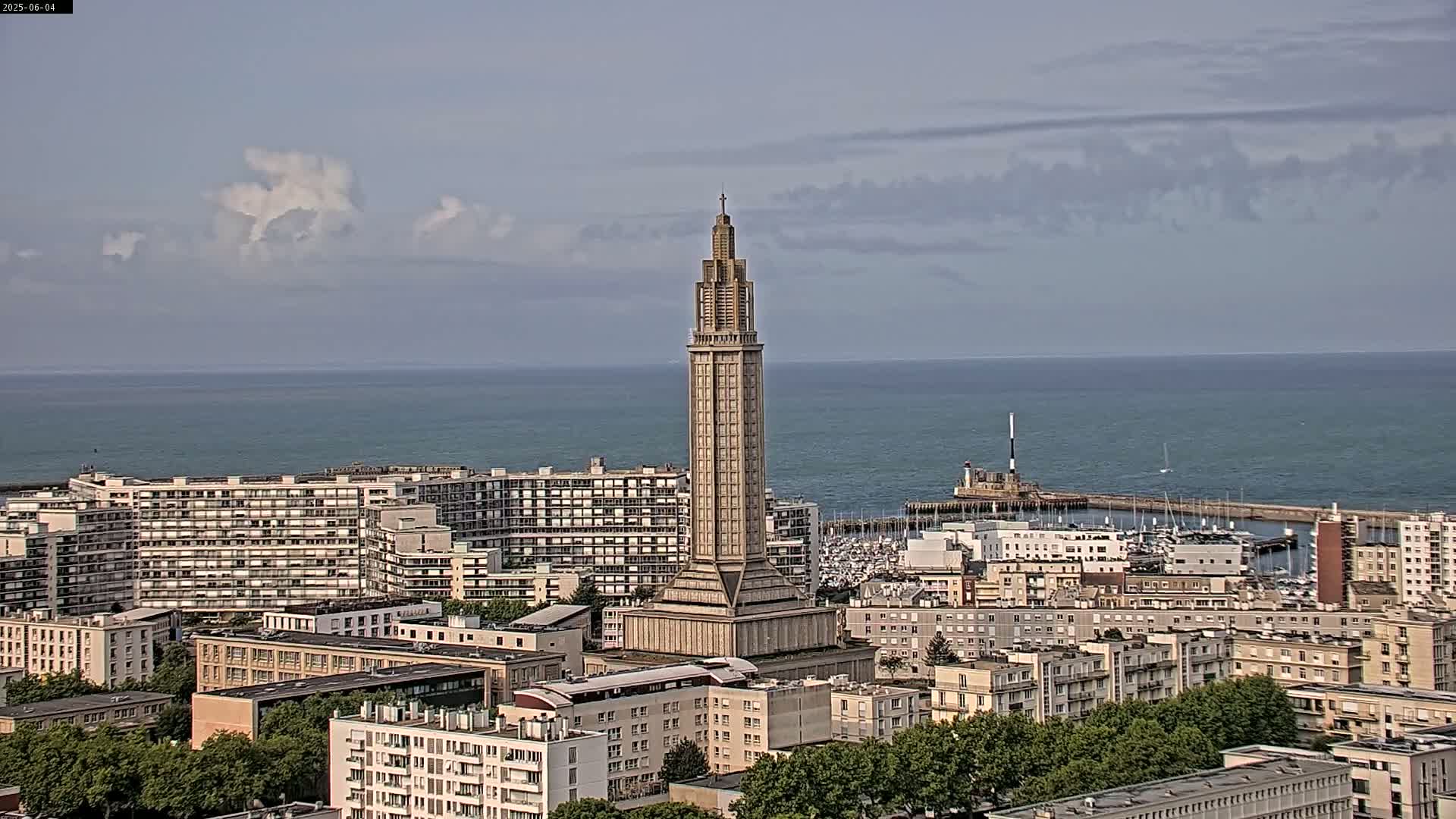 A tall, slender tower stands amidst a city overlooking a calm ocean under a partly cloudy sky.
