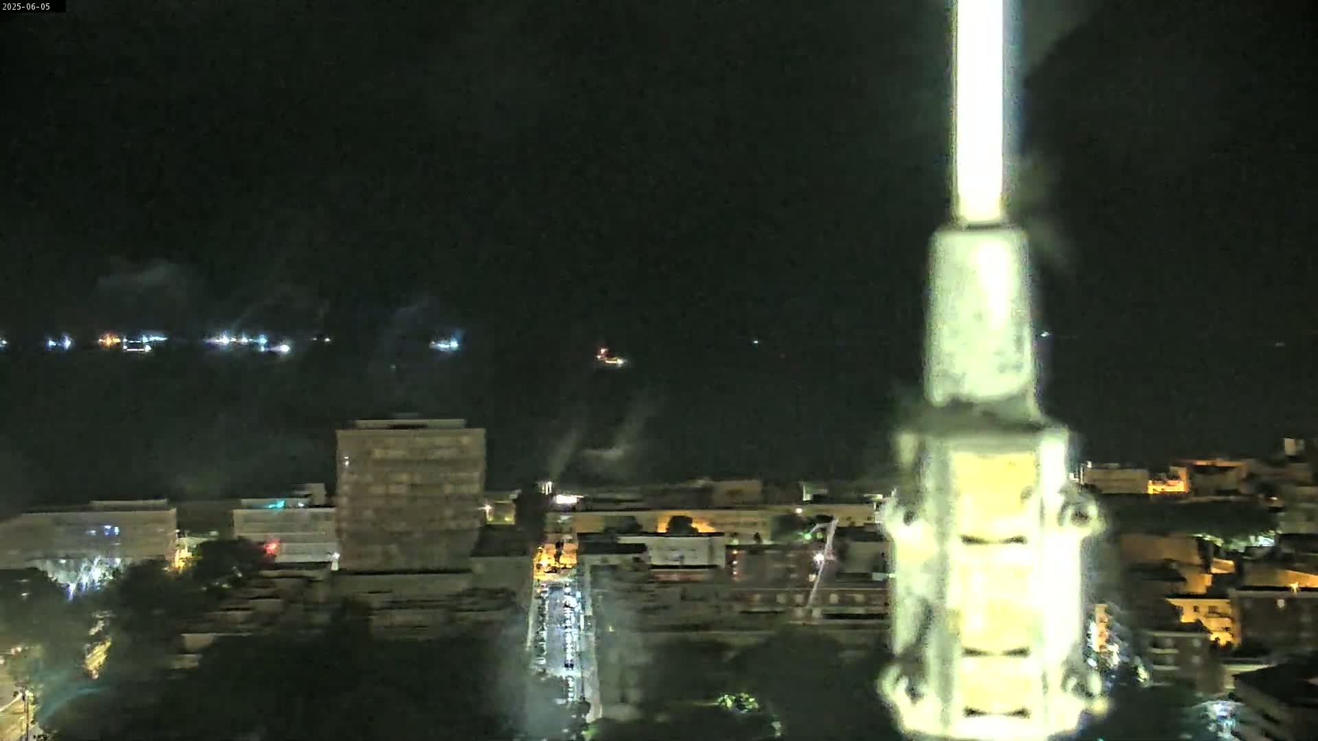A nighttime, high-angle view shows a city skyline next to a dark body of water with lights visible in the distance, a bright light source is prominently featured in the foreground.