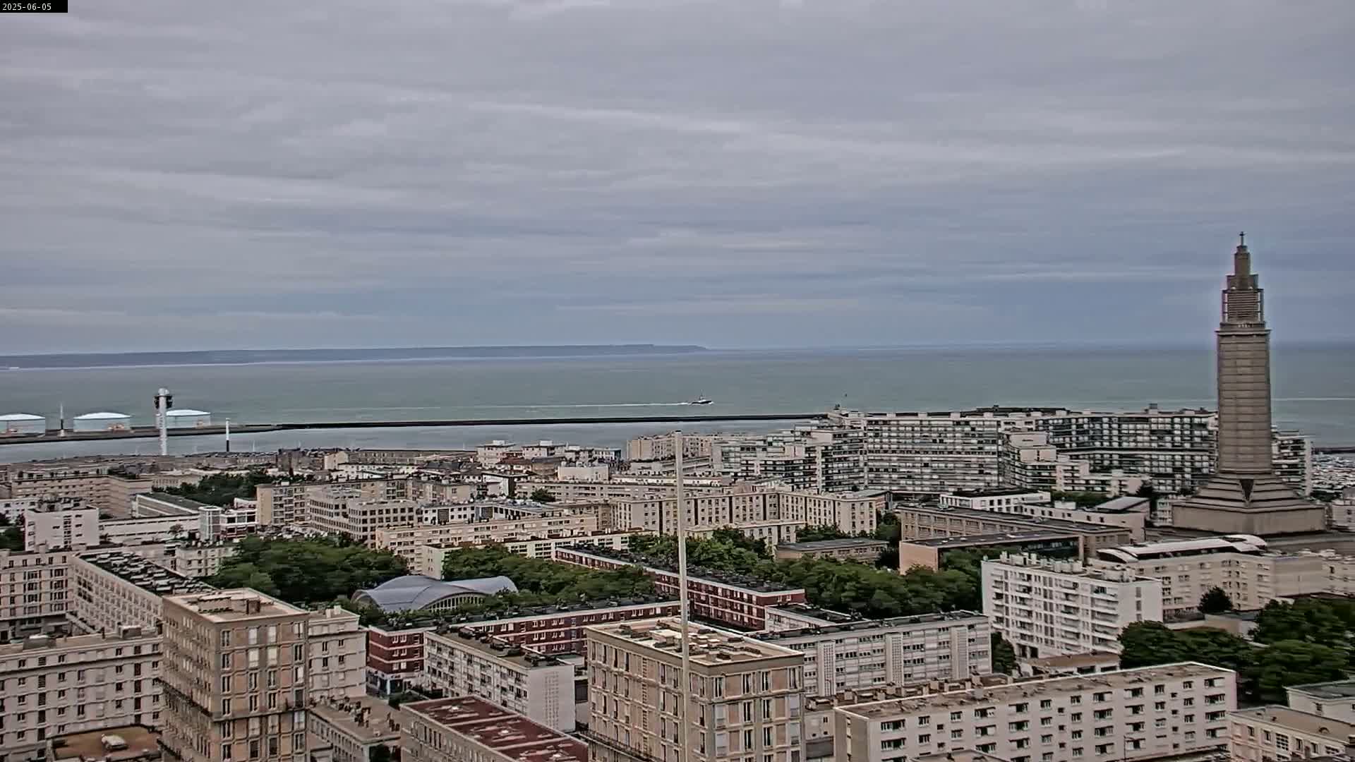 A coastal city under an overcast sky is seen from an elevated viewpoint, showing a tall tower and a large body of water in the distance.