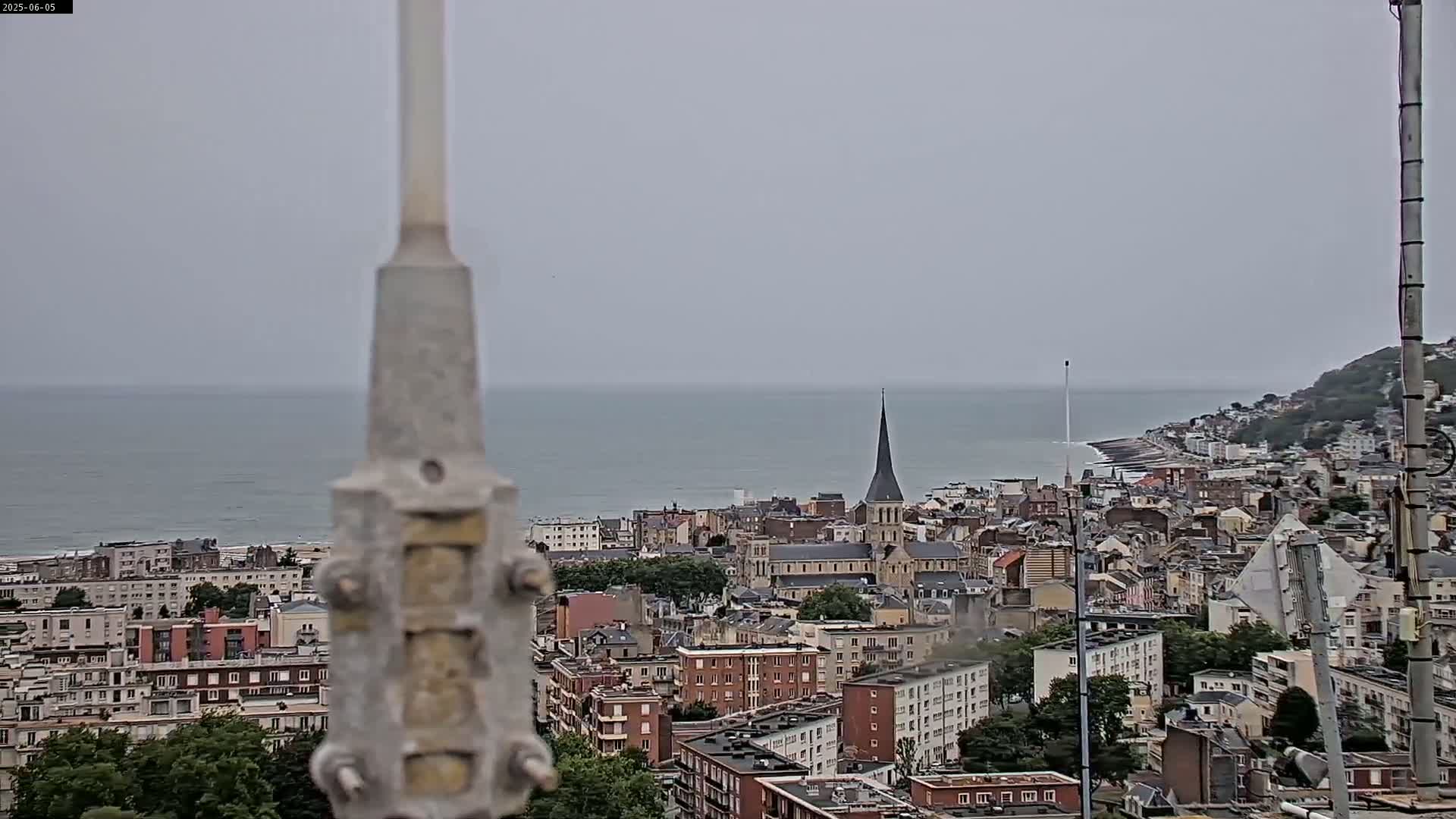 A coastal town with a church spire is visible under an overcast sky, with a calm sea in the background.