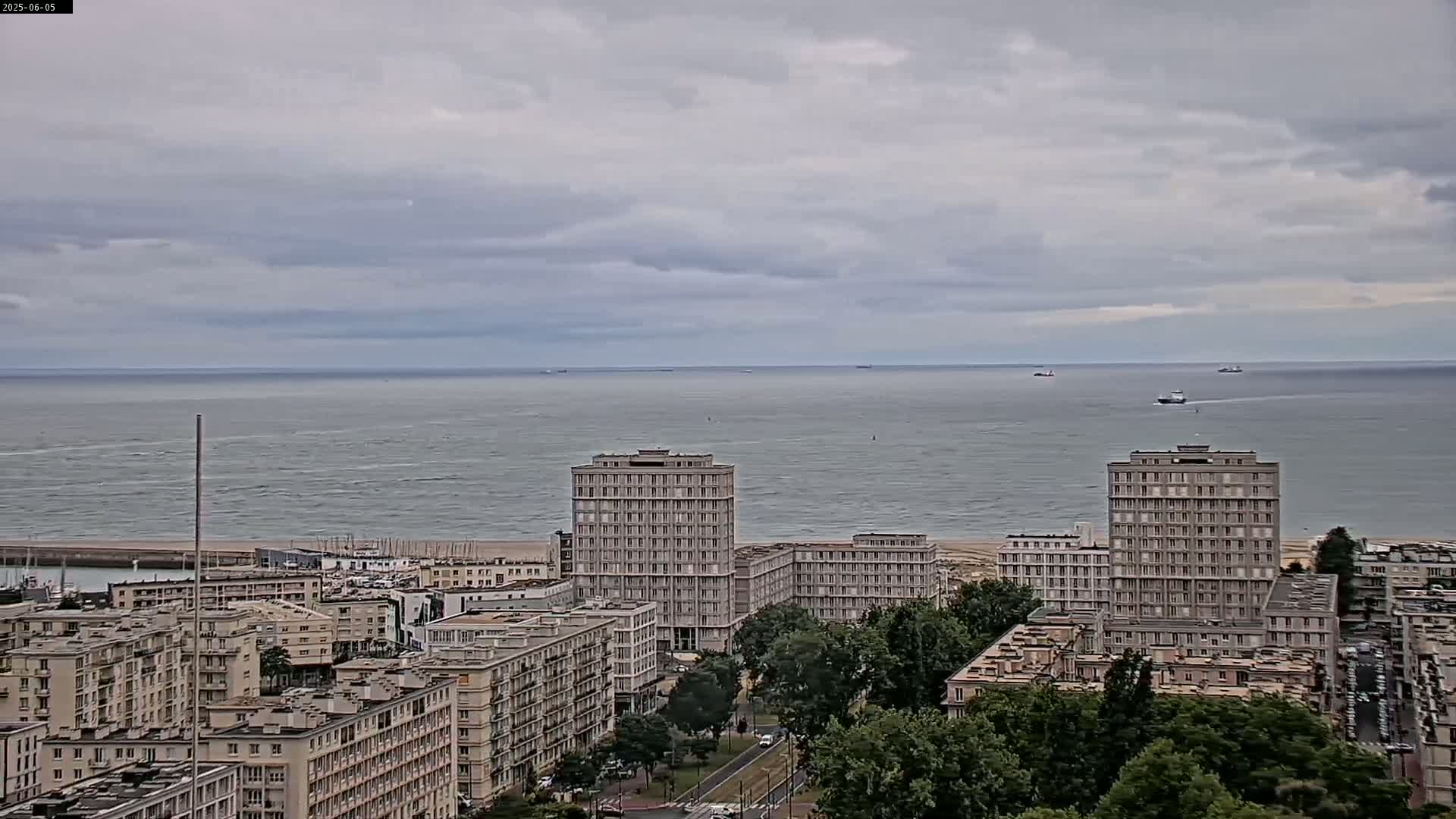 A coastal city with several high-rise buildings is visible under an overcast sky, overlooking a calm sea with a few distant ships.