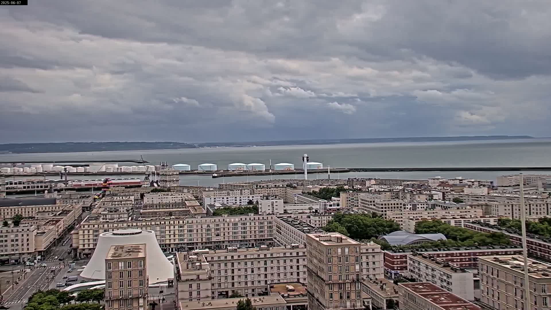 A high-angle view shows a city by a body of water under a cloudy sky, with industrial structures and storage tanks visible near the waterfront.