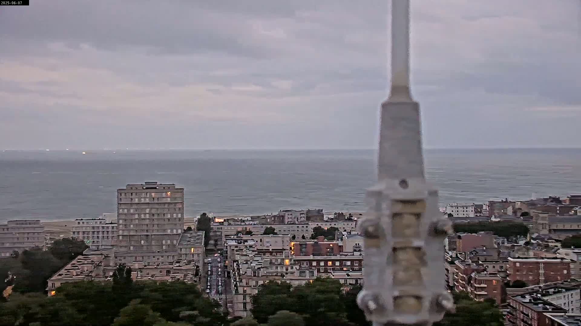 A coastal city with a calm sea under an overcast sky is seen from an elevated vantage point, partially obscured by a tall, slender structure in the foreground.