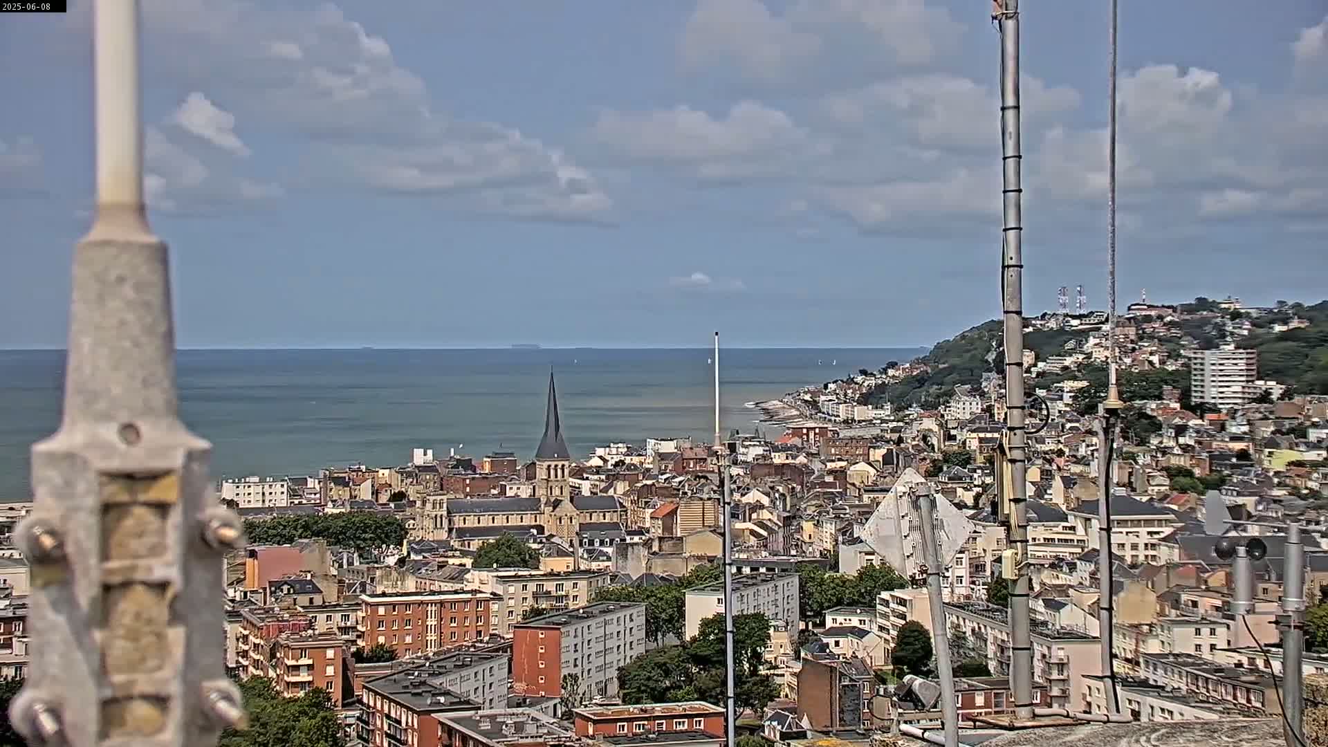 A coastal city with a church spire is visible under a partly cloudy sky, viewed from a high vantage point with antennas in the foreground.
