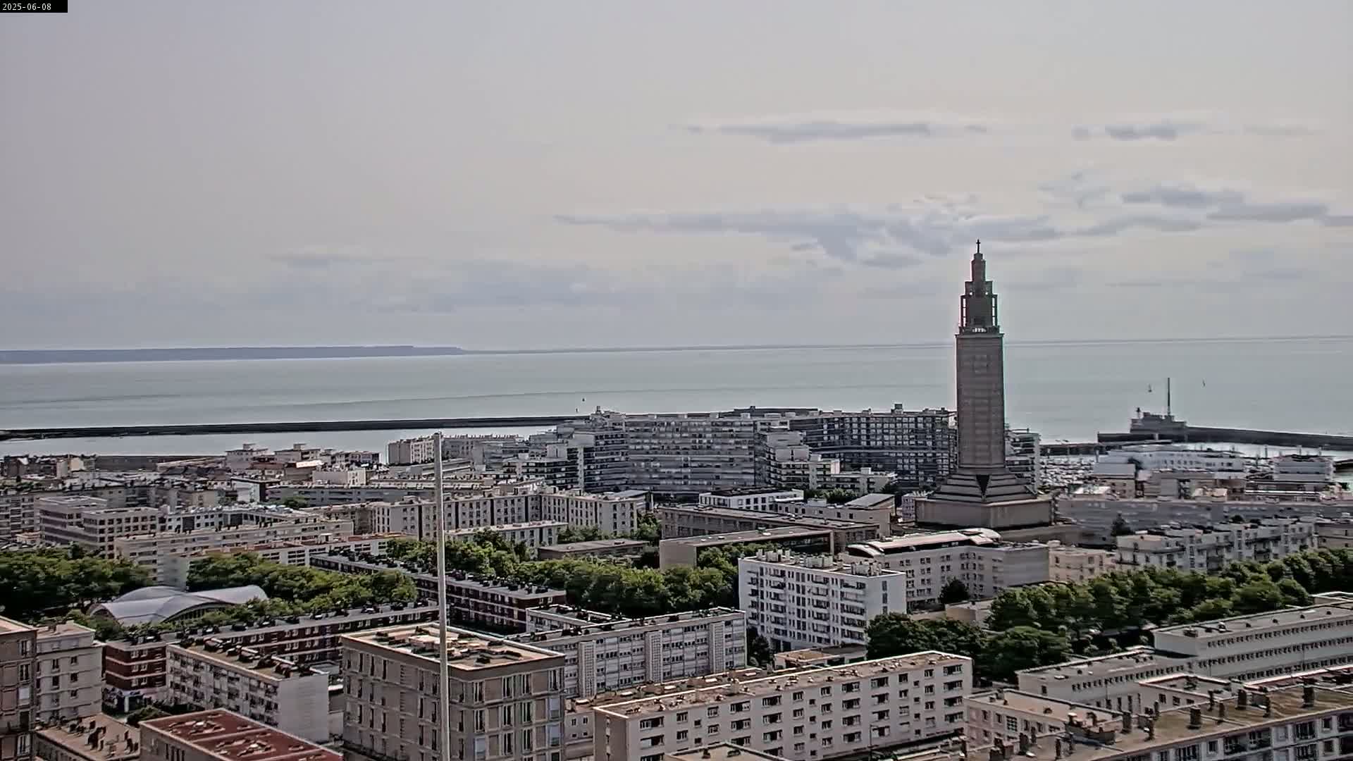 A coastal city with a tall, slender tower is visible under a mostly cloudy sky, overlooking a calm body of water.