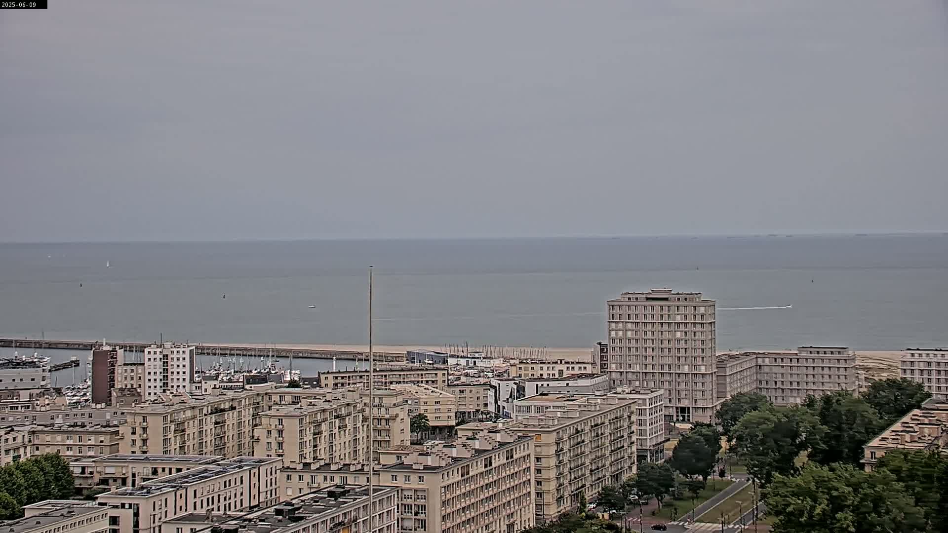 A coastal city with numerous apartment buildings is viewed from above on an overcast day, overlooking a calm sea with a few boats visible.