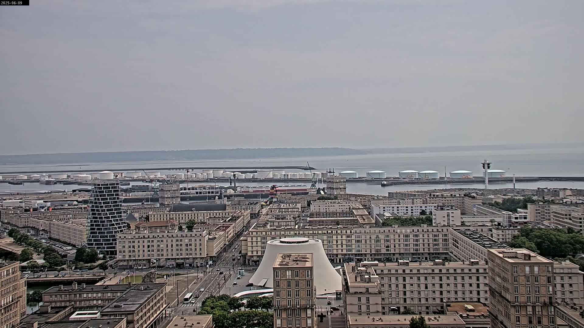 A hazy view of a city harbor with numerous buildings and industrial structures, including large storage tanks, under an overcast sky.