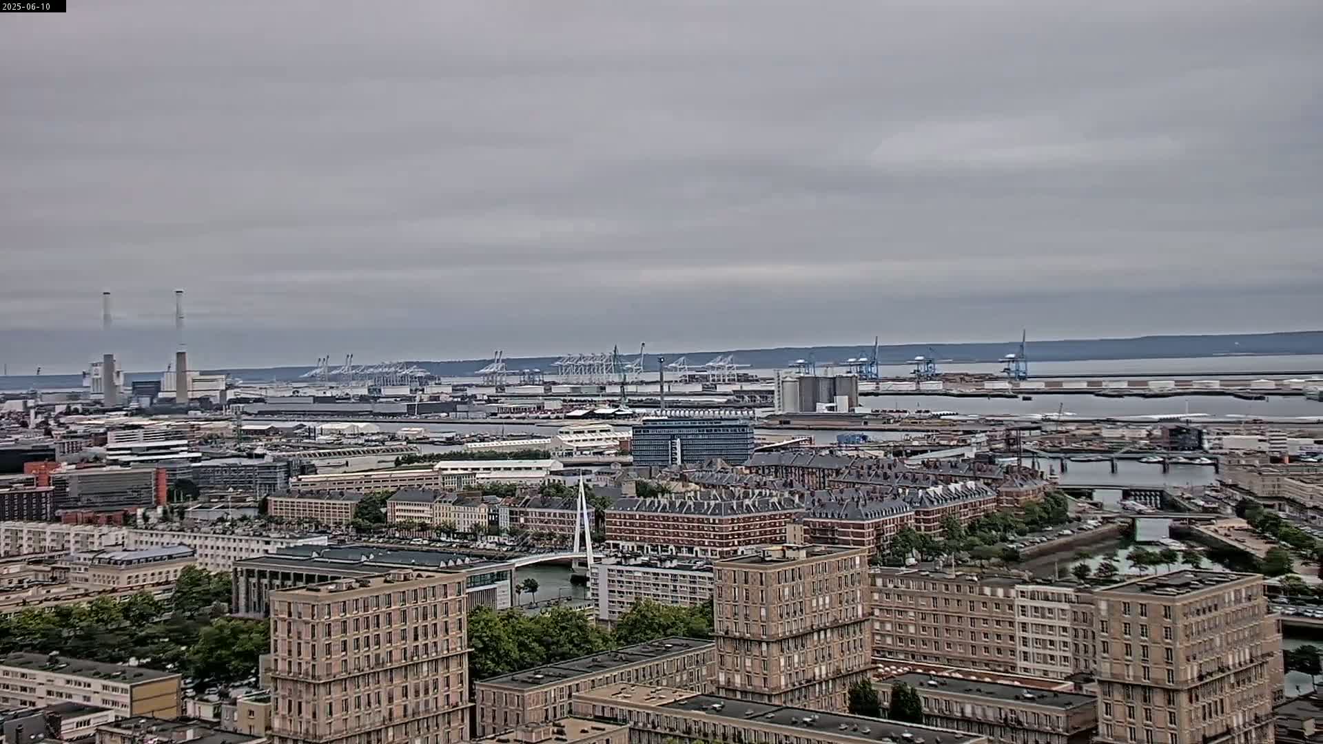 An overcast day reveals a cityscape with numerous buildings, a waterway, and a distant industrial port featuring many cranes under a gray sky.