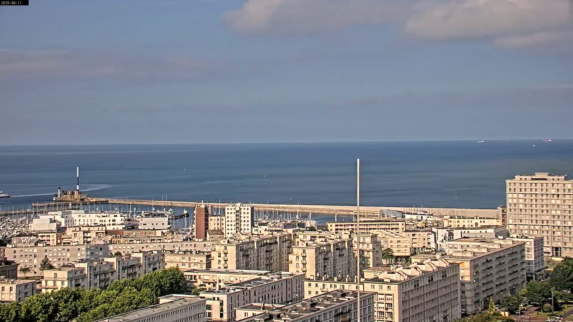 A coastal city with a harbor full of boats is visible under a partly cloudy sky, with a calm ocean stretching to the horizon.