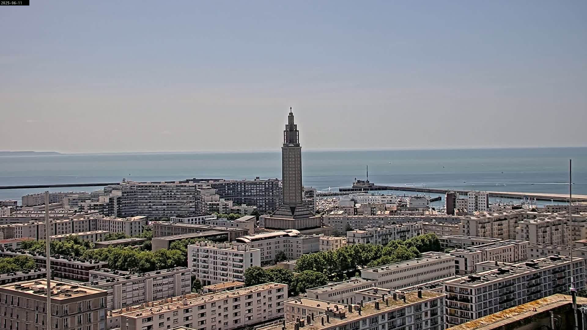A tall, slender tower stands in the center of a city overlooking a calm ocean on a clear, sunny day.