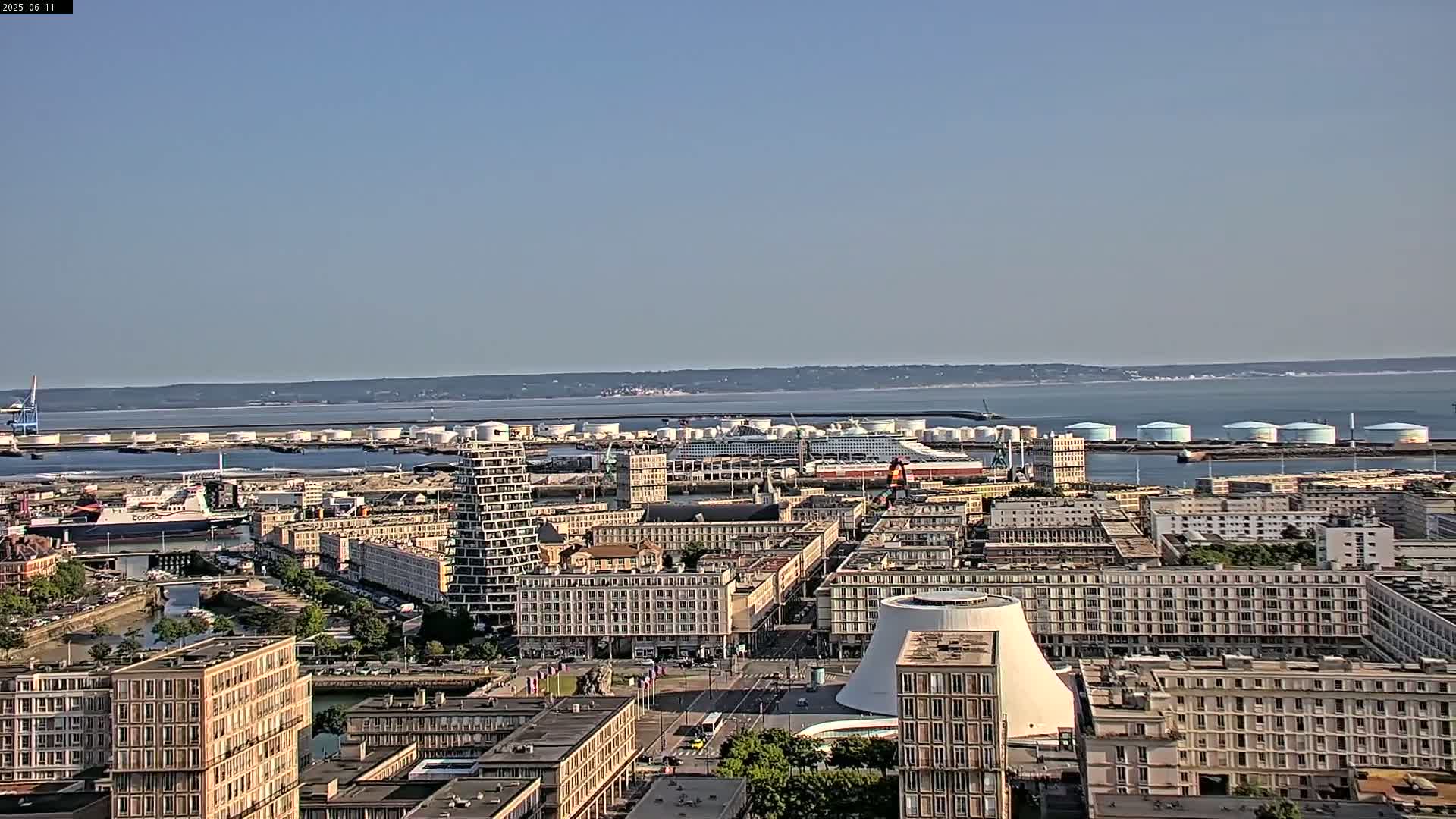 An aerial view of a city harbor on a clear, sunny day shows a cruise ship docked near large storage tanks, with numerous buildings and a waterway visible.