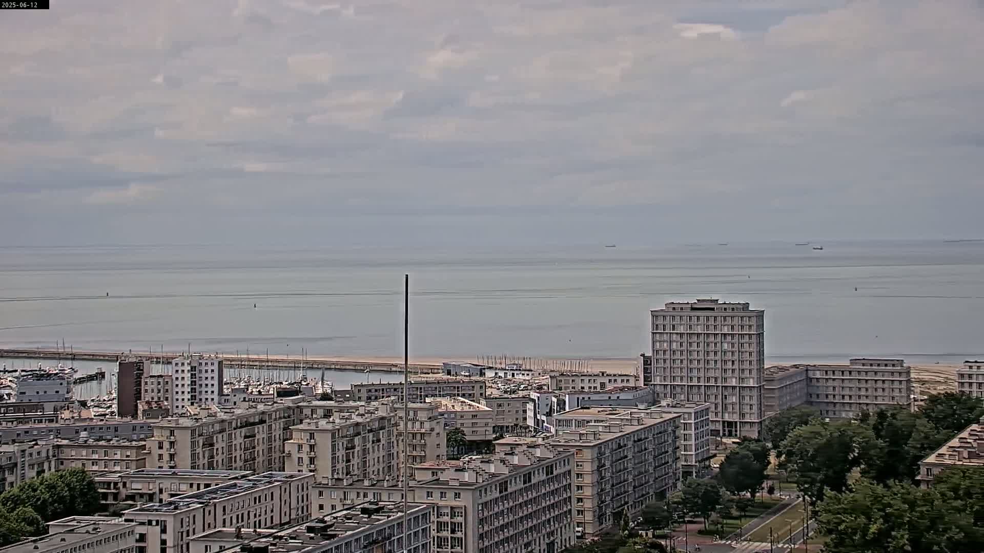 A coastal city with many apartment buildings under a cloudy sky overlooks a calm sea with a few distant ships.