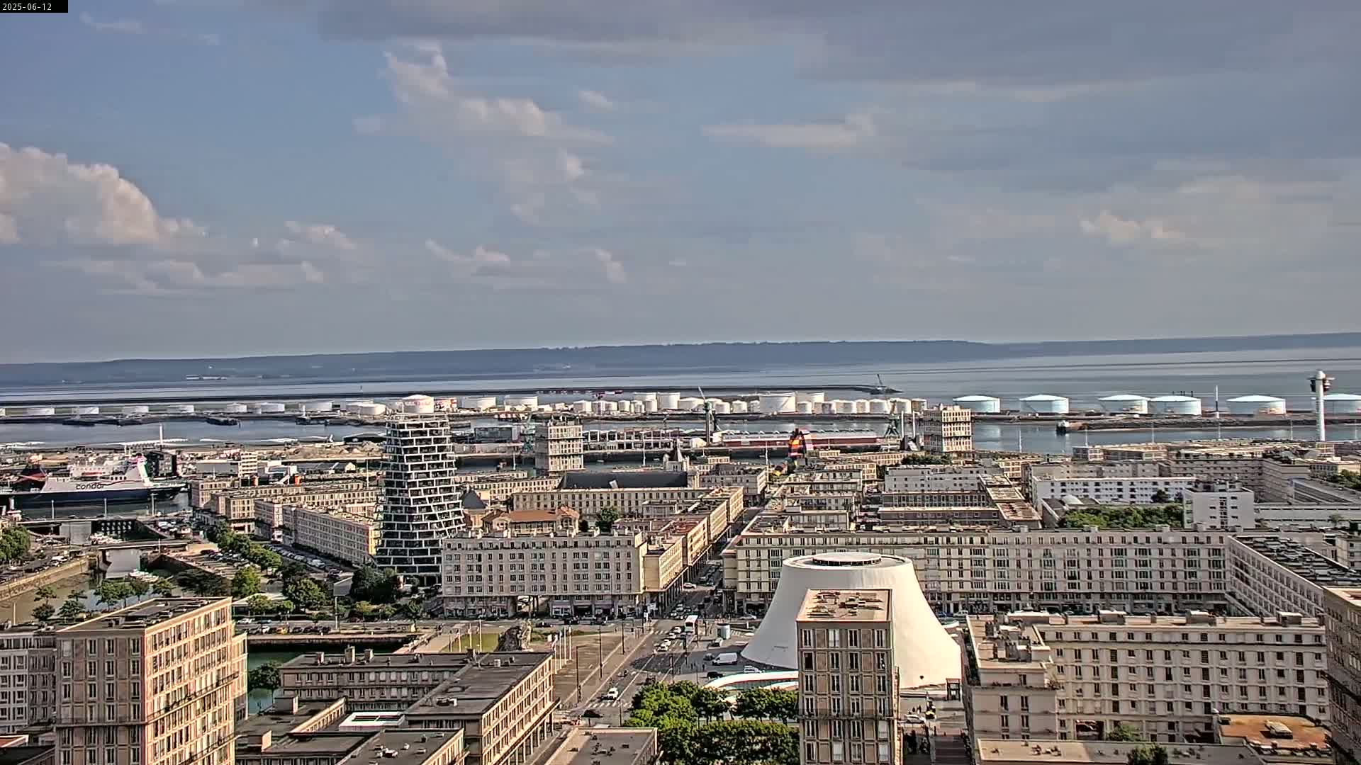 A partly cloudy day reveals a city's waterfront, showcasing a harbor filled with docked ships and industrial storage tanks, all overlooked by numerous apartment buildings.