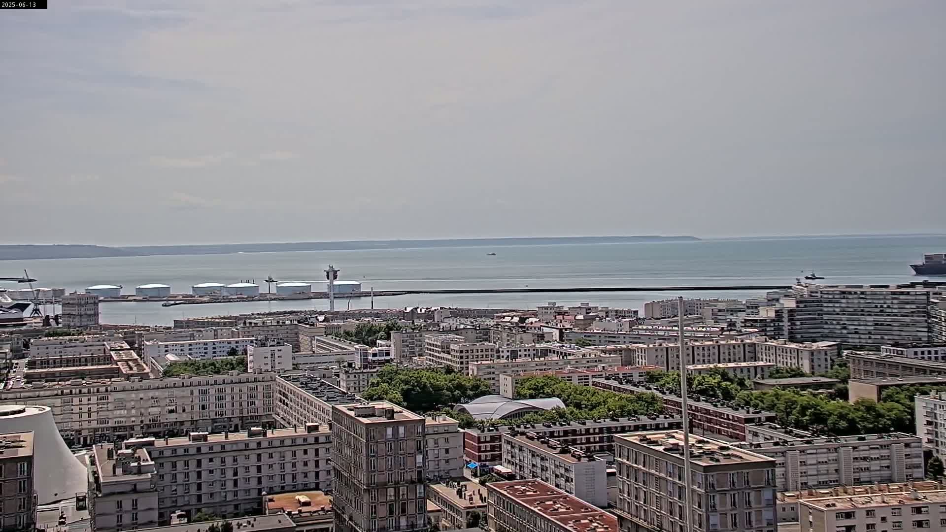 An aerial view shows a city harbor with numerous buildings and storage tanks, next to a calm body of water under a lightly overcast sky.