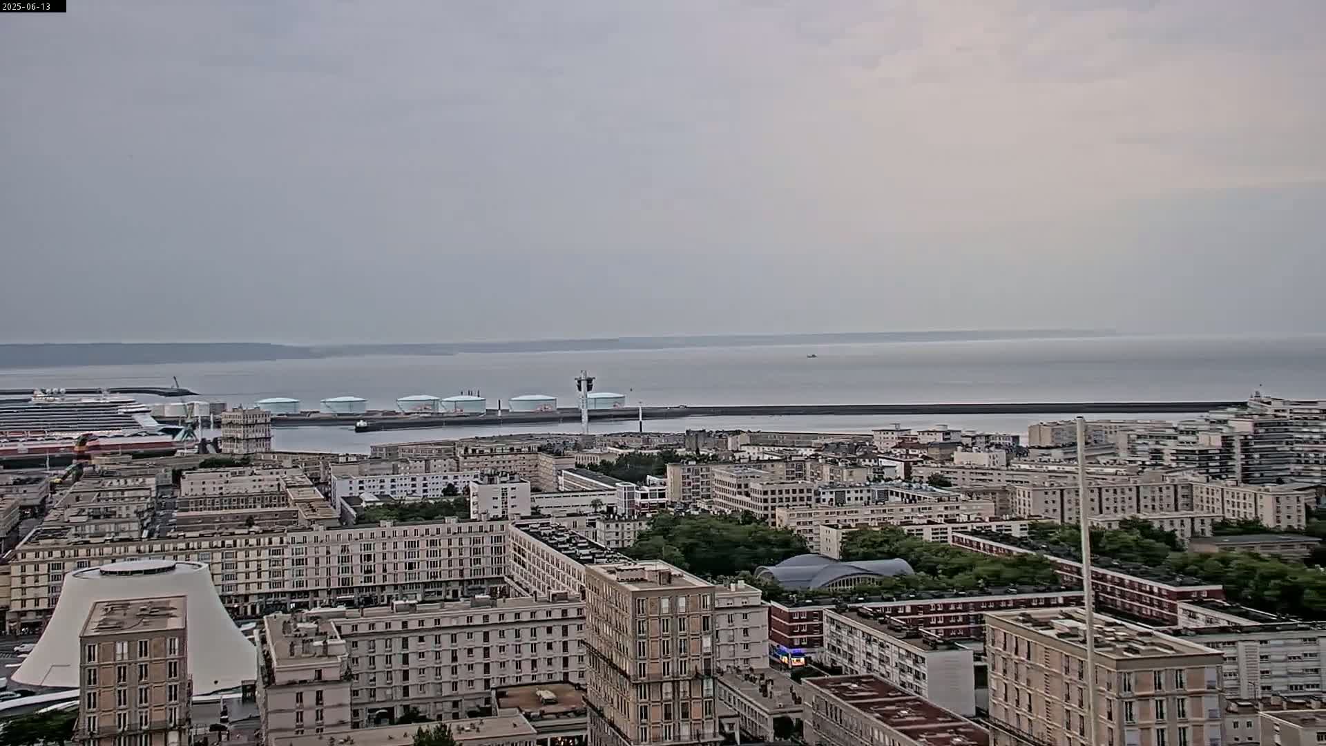 An overcast day reveals a cityscape with numerous buildings, a cruise ship in port, and several large storage tanks near the water's edge, all overlooking a calm body of water.