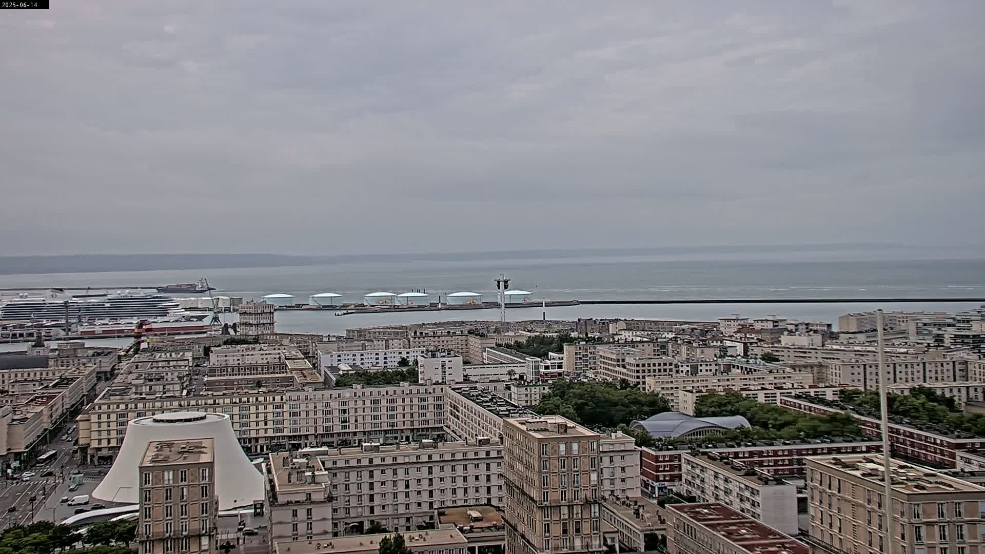 A sprawling cityscape with numerous multi-story buildings extends to a harbor containing a cruise ship and several industrial structures, all under an overcast sky.