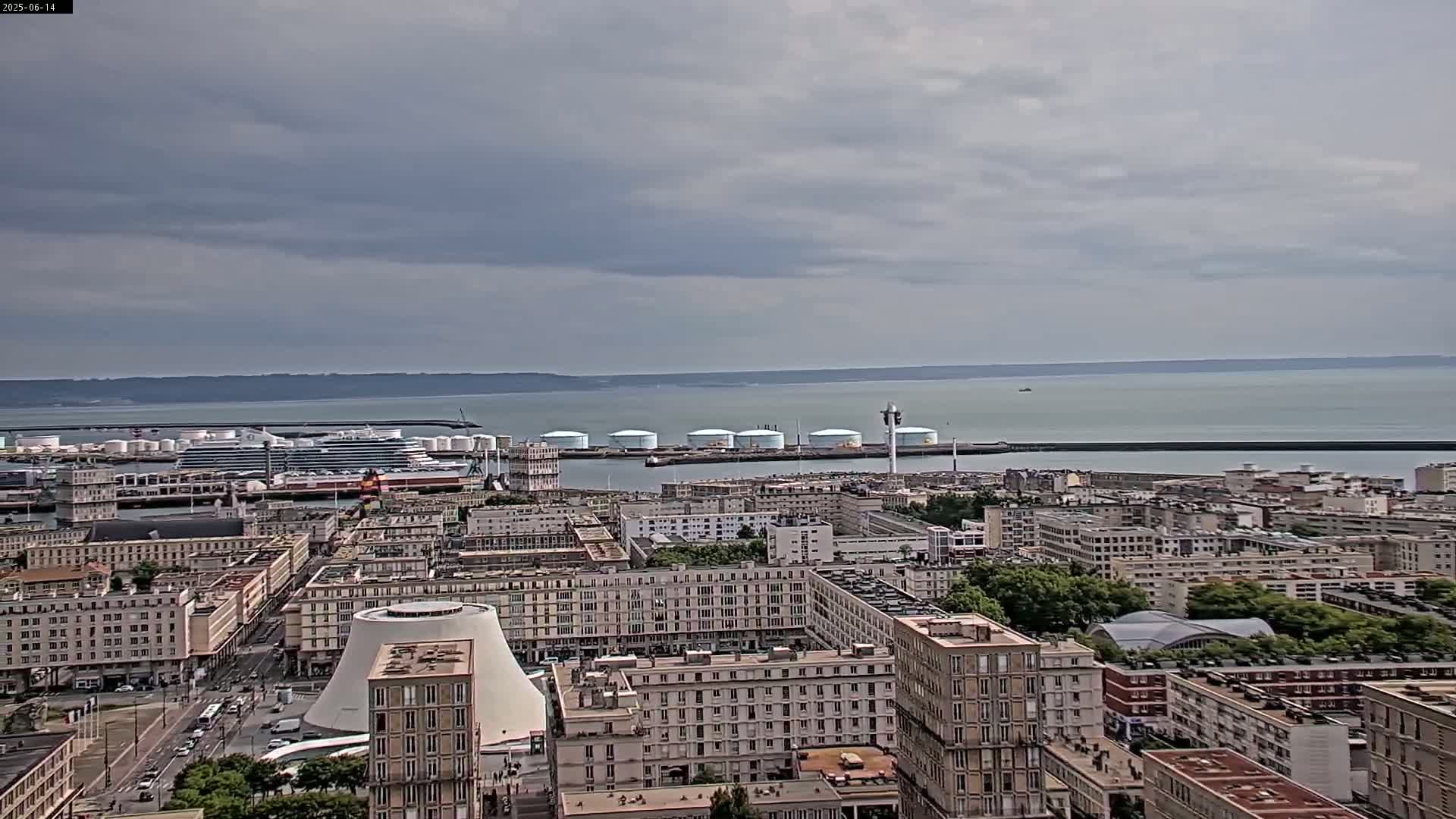 An aerial view of a city harbor on an overcast day shows a large cruise ship docked near several industrial storage tanks, with numerous buildings extending into the distance.