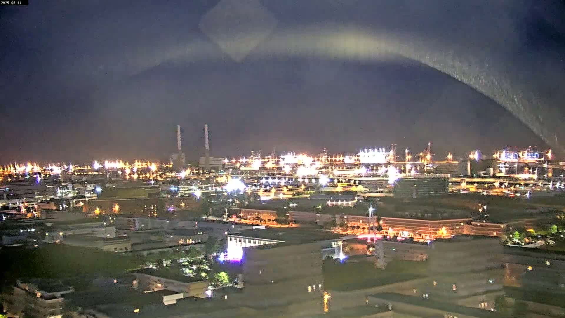 An aerial view of a city harbor on an overcast day shows a large cruise ship docked near several industrial storage tanks, with numerous buildings extending into the distance.