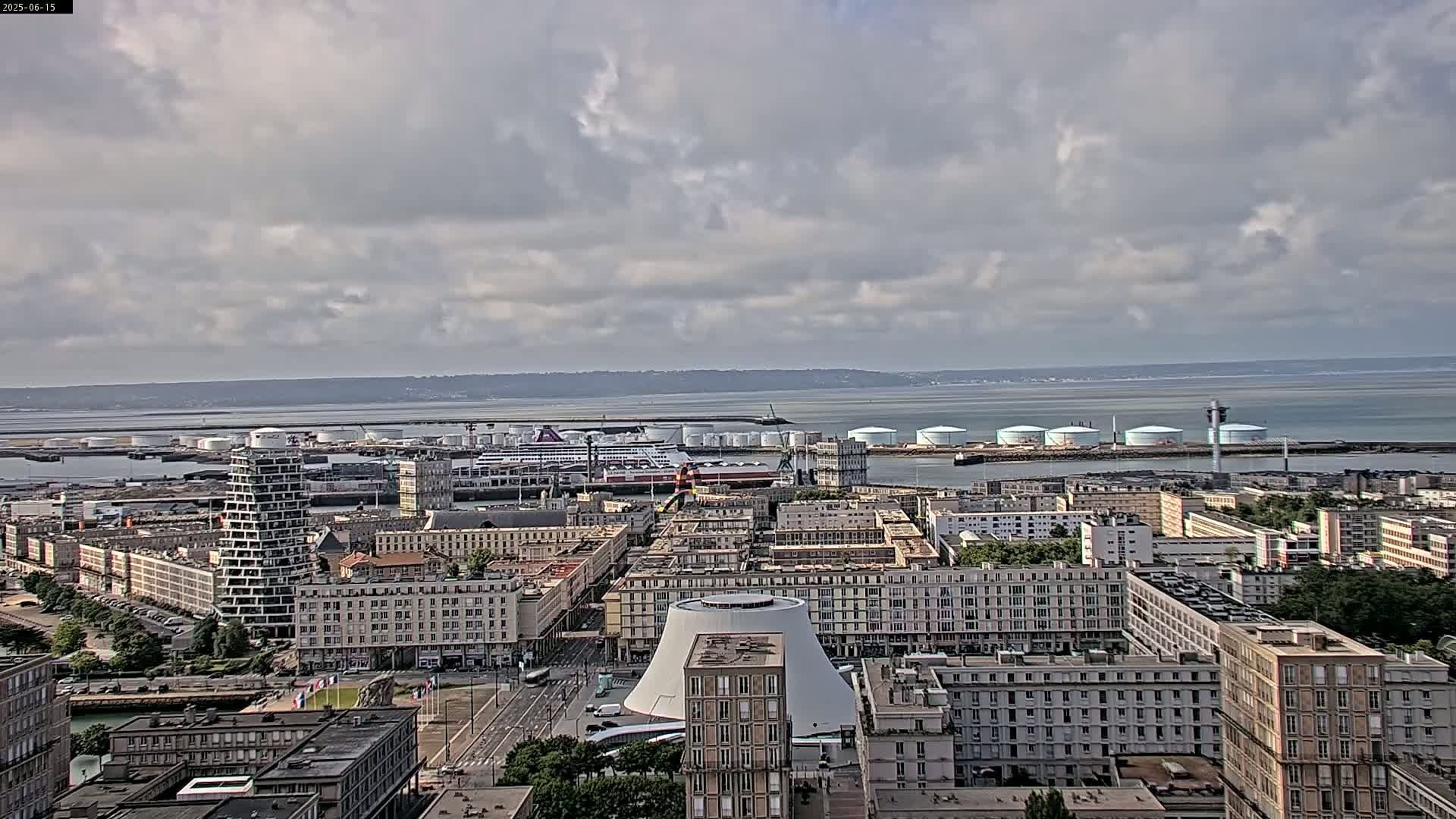 An aerial view of a city harbor on a partly cloudy day shows a large cruise ship docked near several large storage tanks, and numerous buildings extend inland from the port.