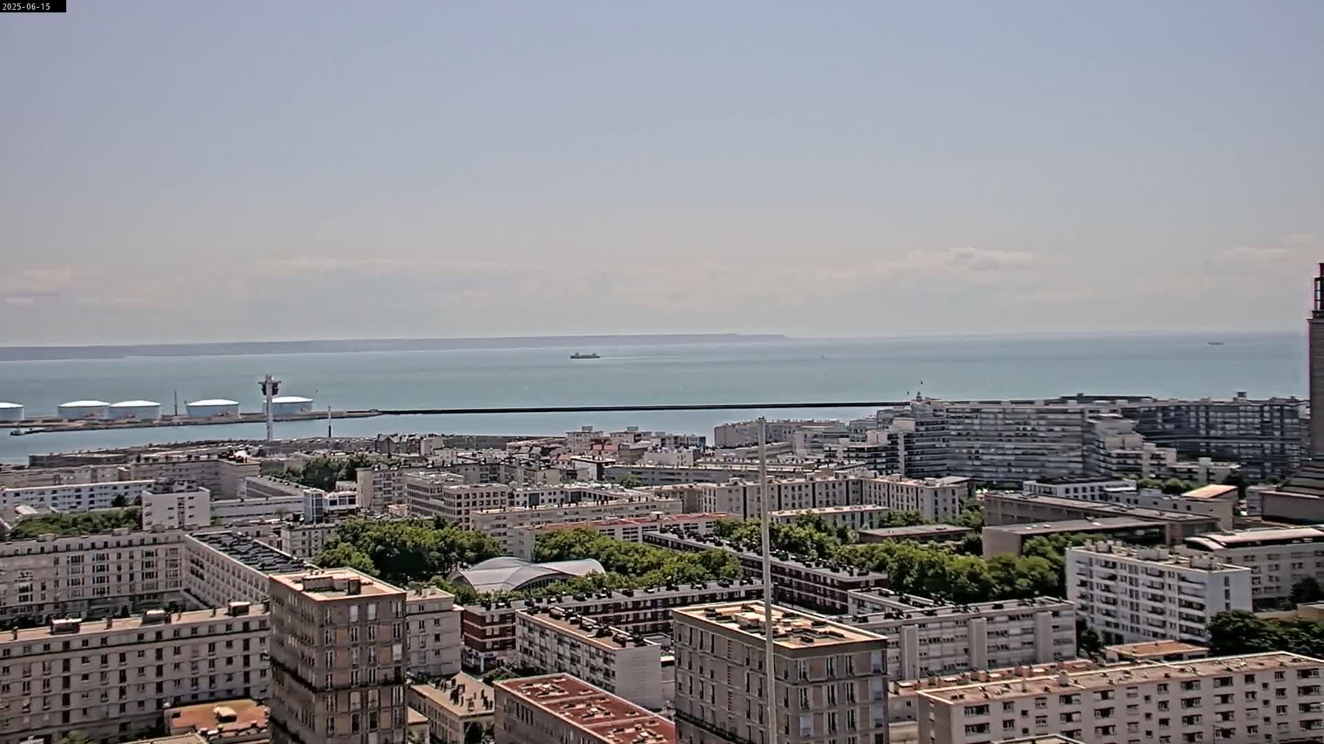 A sprawling cityscape featuring numerous apartment buildings extends to a calm body of water under a mostly clear sky.
