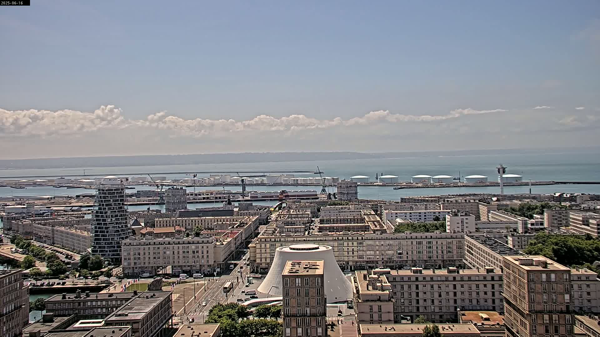 A city harbor with numerous buildings and oil storage tanks is visible under a partly cloudy sky.