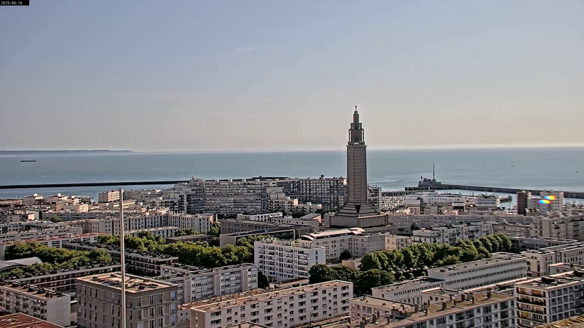 A coastal city with a tall, slender tower is visible under a clear, sunny sky, overlooking a calm sea.