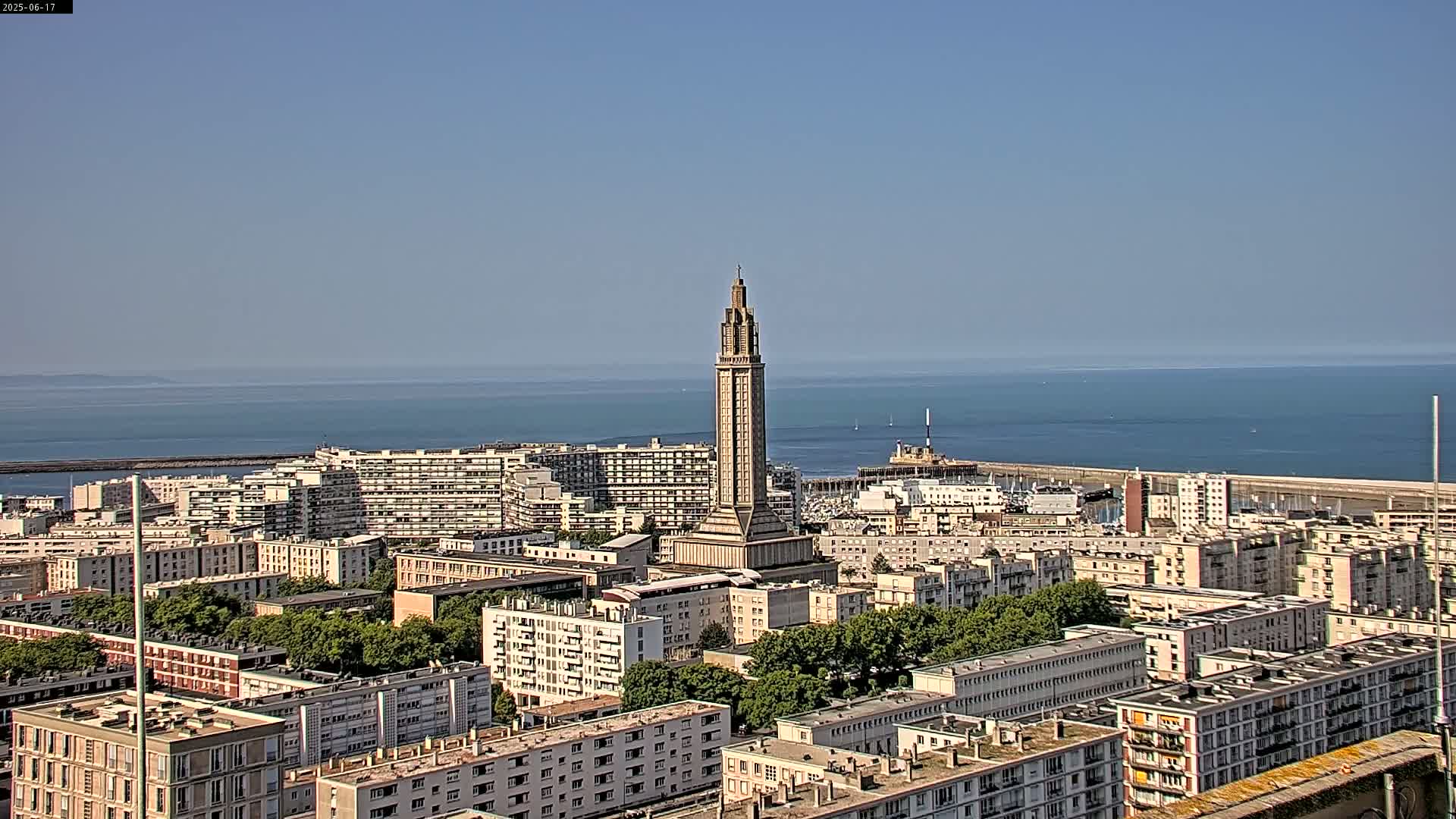 A tall, slender tower stands in the center of a coastal city under a clear, sunny sky, overlooking a calm ocean.